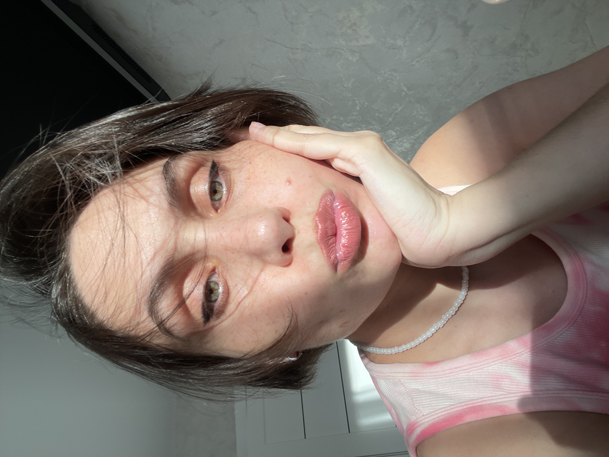A close-up, angled selfie shows a young woman with short, dark hair and light skin. Her eyes are light brown, and she wears dark eyeliner. She has freckles on her face. Her right hand is resting on her cheek. She wears a pink and white tie-dye tank top and a white beaded necklace. The background shows a grey wall with a window. The location is not provided in the image.