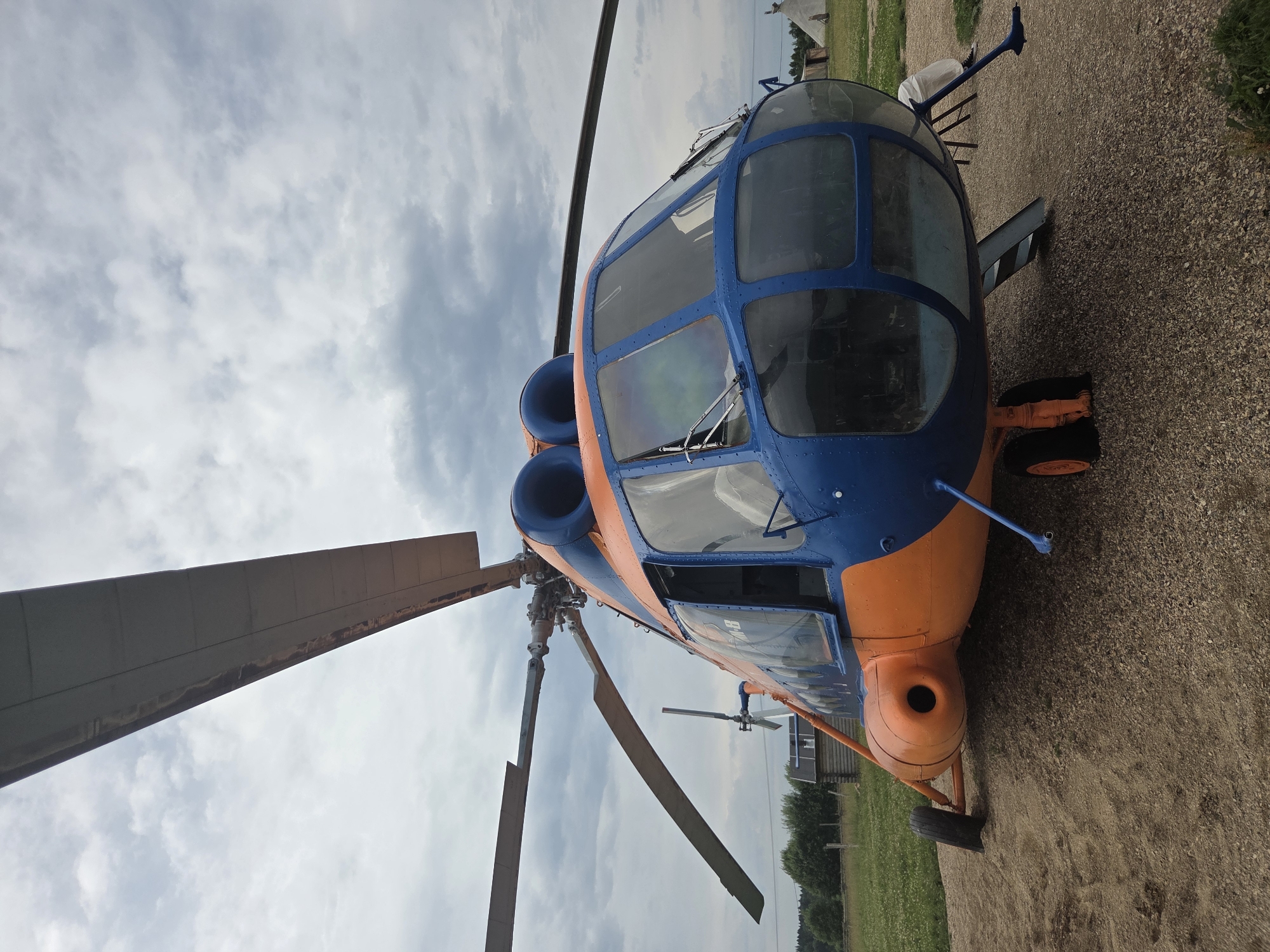 This is a low-angle, full-frame photograph of a blue and orange helicopter parked on a gravel surface. The helicopter is positioned in the center of the frame and occupies the majority of the image. The front of the helicopter, with its distinctive blue cockpit and large windows, faces the viewer. Two large rotor blades extend upwards and outwards from the top of the helicopter. The helicopter's body is predominantly orange, with blue accents around the cockpit and engine cowlings. One of the cabin doors is open. The helicopter is resting on three landing gear wheels, one of which is partially obscured by the gravel. In the background, there is a green grassy field with trees and some small wooden structures, including what appears to be a small log cabin and a conical tent-like structure. The sky is overcast with dramatic grey and white clouds. The ground beneath the helicopter is composed of loose gravel and dirt. The overall scene suggests an outdoor display or static exhibition of the aircraft, likely in a rural setting. The location in Mashino, Russia is not visually confirmed by the image itself, but assumed from the prompt.