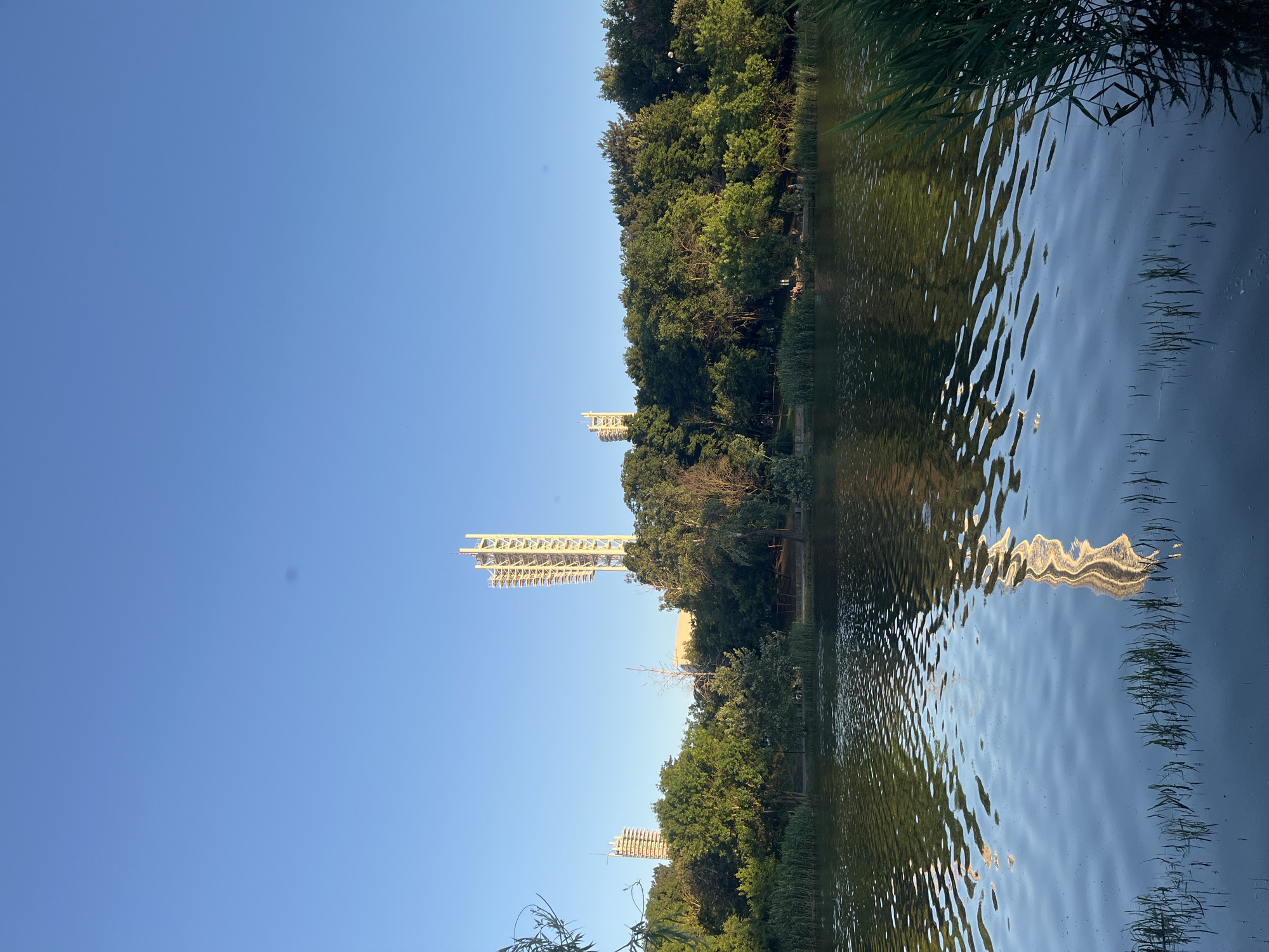 This image captures a serene scene at the Solnechny Ostrov (Sunny Island) Park in Krasnodar, Russia. In the foreground, a calm body of water reflects the clear blue sky and the surrounding greenery. Tall reeds grow along the water's edge on the right side of the frame. In the midground, a dense line of trees forms a lush backdrop, with some taller buildings and a distinctive tall, white structure with a lattice-like design visible behind the foliage. The tall structure appears to be a sports stadium or a similar large-scale facility, possibly with lighting towers. The overall atmosphere is peaceful, suggesting a late afternoon or early evening setting with soft natural light. A few distant figures can be seen among the trees, indicating the presence of park visitors.