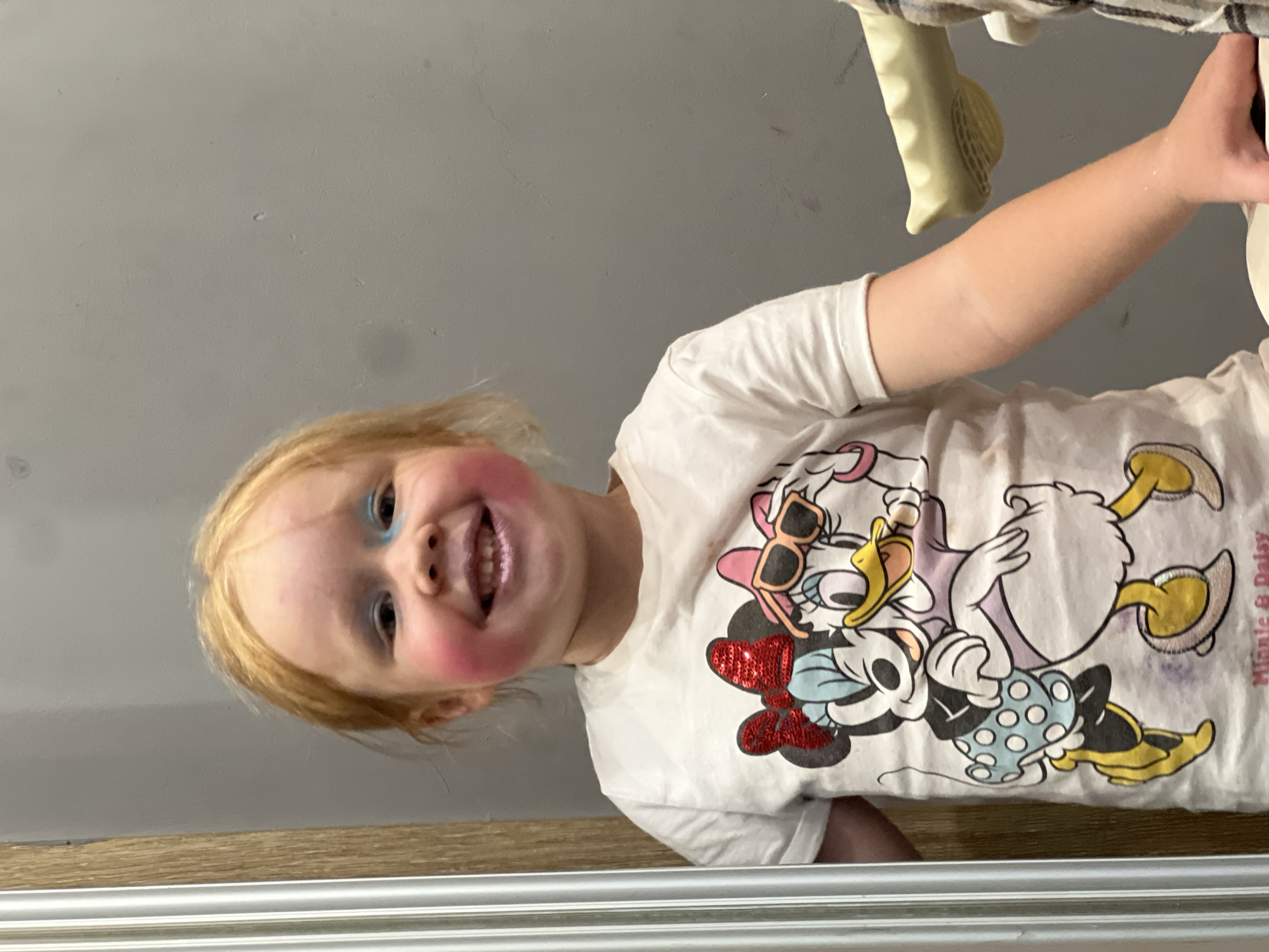 A young child with red hair and makeup applied to their face is standing in front of a gray wall. The child is wearing a white t-shirt with an image of Minnie Mouse and Daisy Duck on it. The child's face is smudged with blue and pink makeup, and their lips have a purple shimmer. The child is smiling and looking towards the camera. The background appears to be an interior space, possibly a bathroom or dressing room, with a mirrored surface visible on the left side of the image and a portion of a light-colored object on the right. The location is not specified in the image or the prompt.