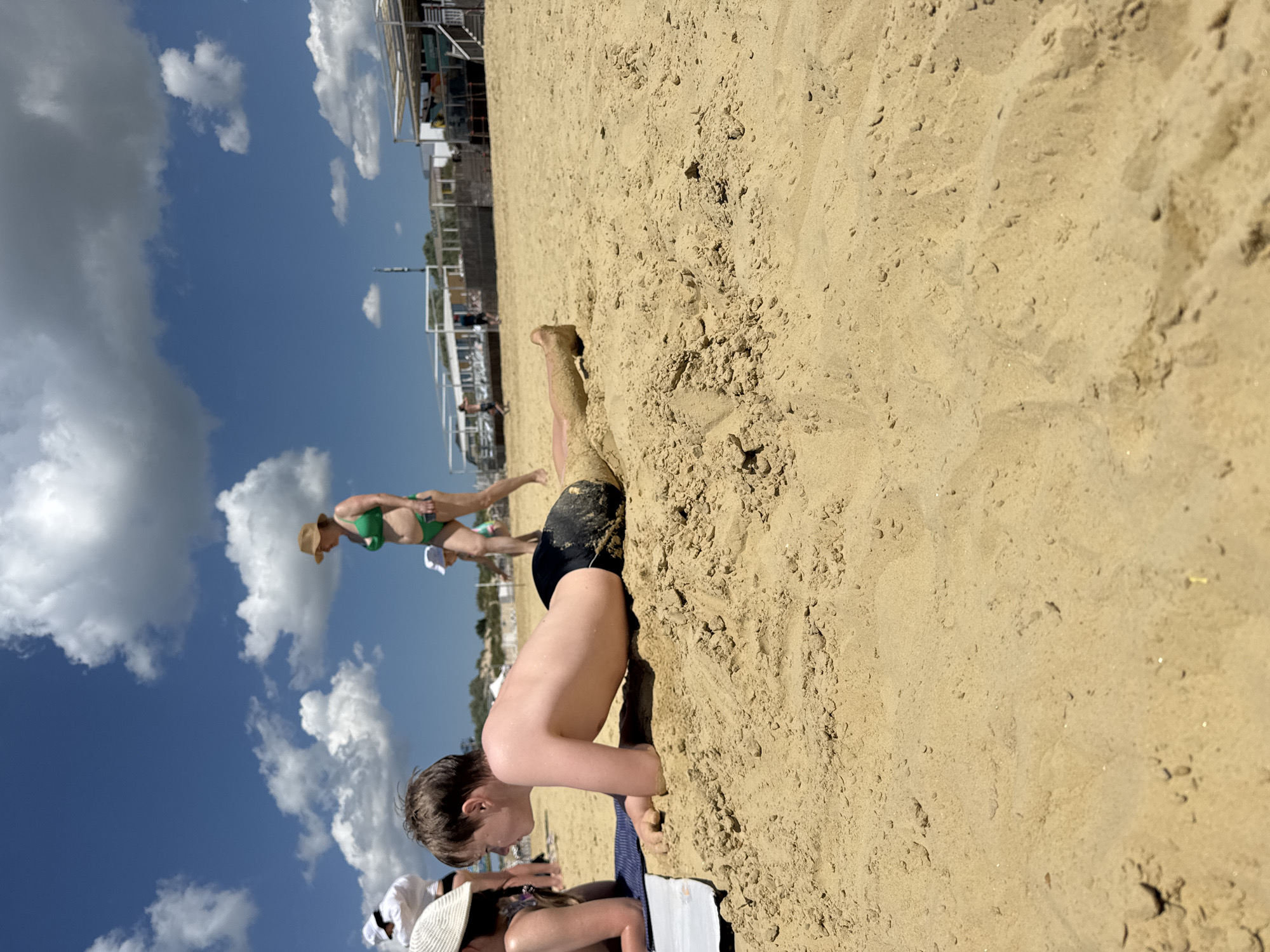 The media file depicts a sunny day at a sandy beach in Anapa, Russia. The main focus is a young boy buried in the sand up to his chest, with only his head and arms visible. He appears to be enjoying himself, as indicated by his smile. In the background, other beachgoers are visible, including a woman in a green bikini and a straw hat, and a person in green swimming trunks. There are also beach umbrellas and some structures in the distance. The sky is blue with scattered white clouds, suggesting fair weather. The sand is a light golden color, typical of a beach environment. The low angle of the sun suggests it is daytime, possibly late morning or afternoon. There is no visible text in the media file.