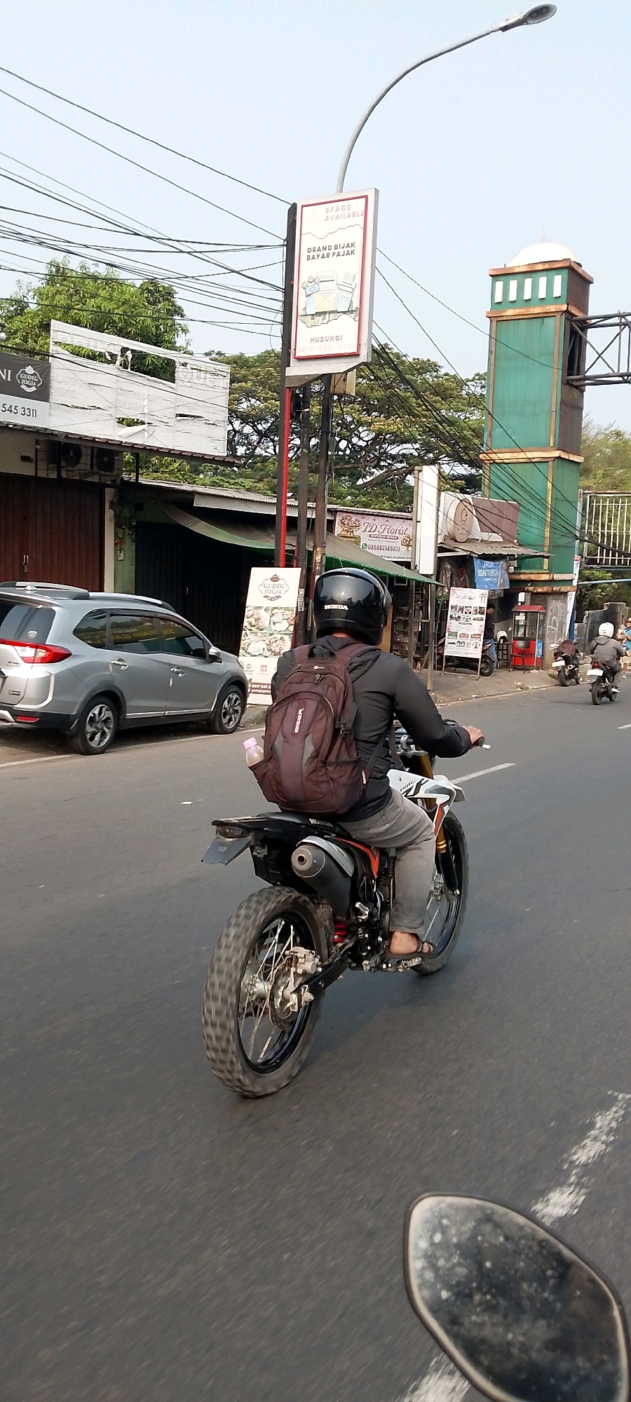 A low-angle, full-shot photograph captures a man riding a dirt bike on a paved road in Bekasi, Indonesia. The rider, wearing a black helmet and a dark jacket, has a maroon backpack with a water bottle in its side pocket. He is wearing grey pants and brown flip-flops. The dirt bike is black, white, and orange. In the background, a silver SUV is parked on the left side of the road. A green and brown tower with a white dome is visible in the far background, along with a street light and several power lines. Various signs are mounted on poles, including one advertising "SPACE AVAILABLE" and "ORANG BIJAK BAYAR PAJAK". Another sign reads "GUDEG JOGJA". The sky is clear and bright. The road is asphalt and has white lane markings. The overall scene appears to be a busy street in a commercial or mixed-use area.