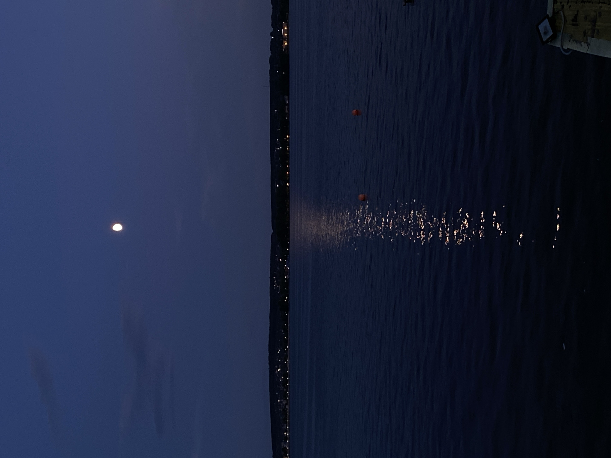 This image depicts a twilight scene at Votkinsk, Russia, likely overlooking a body of water. The sky is a deep, dusky blue, with the moon faintly visible as a bright, circular orb. The left half of the image is dominated by this expansive sky.
The right half of the image is filled with dark, rippling water. Along the distant shoreline, a line of lights from a settlement twinkles, suggesting a town or village. Two orange buoys are visible on the water's surface, floating some distance apart. A shimmering reflection of light, possibly from the moon or distant lights, dances on the water in the lower portion of the frame. In the upper right corner, a small section of a wooden dock or pier is visible, with a dark rectangular object mounted on it, possibly a small light fixture or sign.
The overall mood is tranquil and serene, indicative of early evening or late dusk. The faint lights and the moon suggest a peaceful atmosphere. There is no visible text in the image.