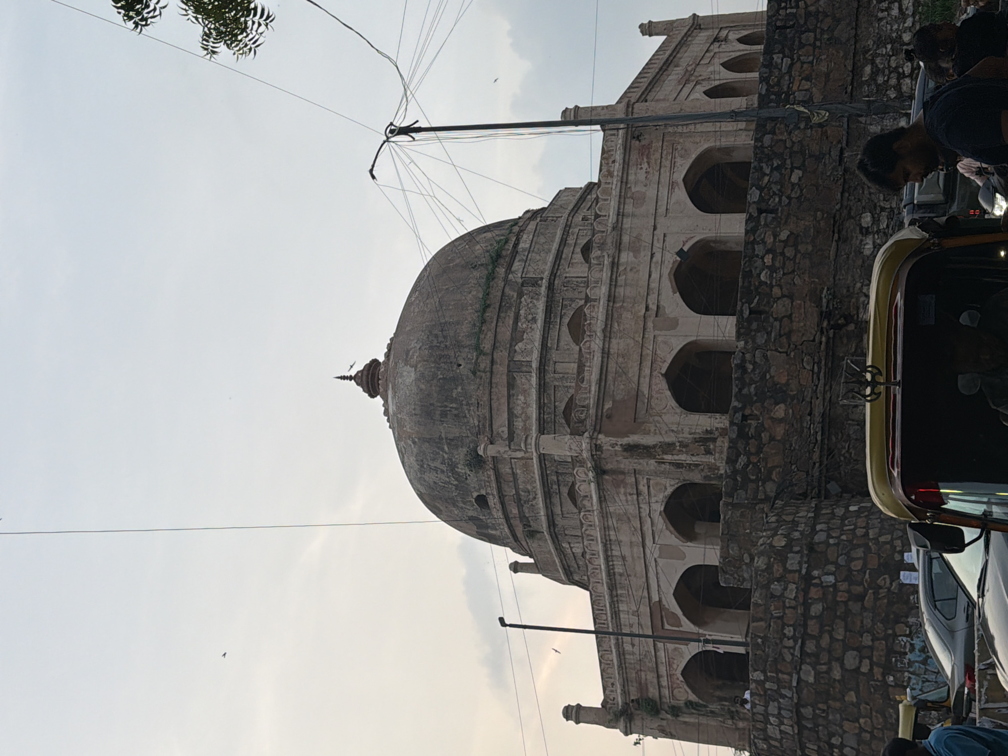 This image shows a dome-shaped building, possibly a tomb, located in Delhi, India. The structure is made of stone and features multiple arched windows and decorative elements. It is surrounded by a stone wall and some greenery. In the foreground, there is traffic with a few vehicles, including an auto-rickshaw. People are visible near the vehicles and on top of the wall of the building. The sky is overcast with some clouds.