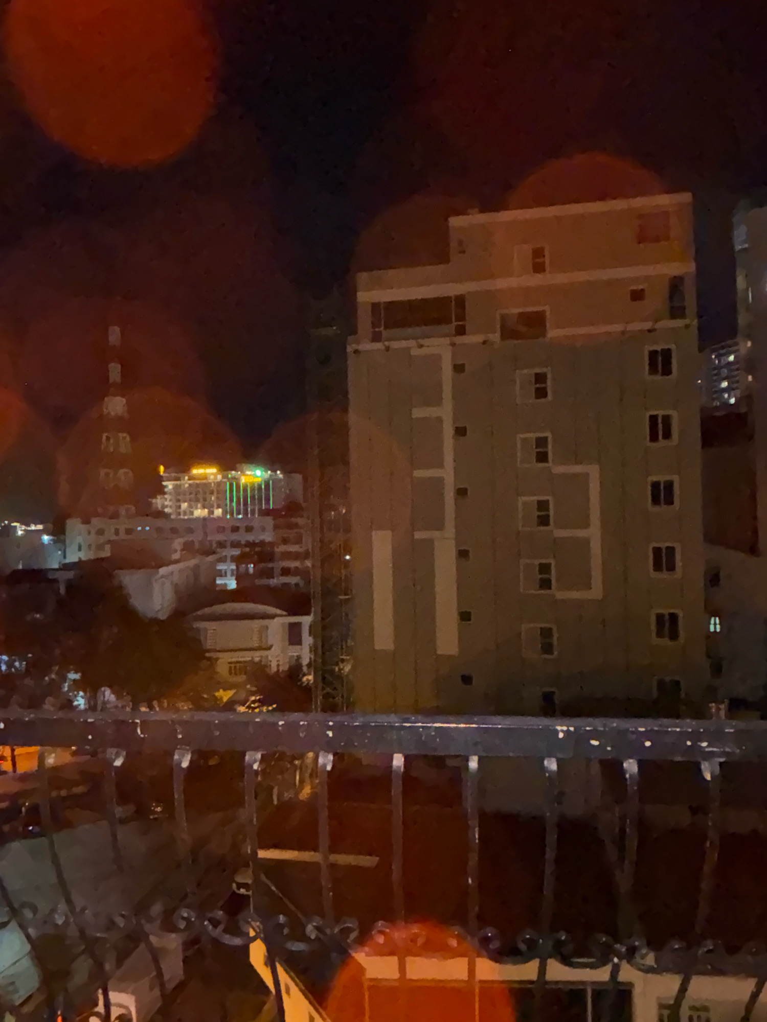 The image depicts a nighttime view of a city skyline, likely in Nha Trang, Vietnam. In the foreground, a dark metal balcony railing is visible. Beyond the railing, several buildings of varying heights are illuminated.
A prominent light-colored, multi-story building with numerous windows occupies the central portion of the frame. It features a grey facade with white trim around the windows and geometric patterns. To the left of this building, a taller structure with a tower-like element is visible, adorned with bright green lights and a sign that appears to read "NHA TRANG". Further in the background, other buildings and structures can be seen, including what appears to be a construction crane to the right of the central building. The overall scene is characterized by artificial lighting against a dark night sky, with some blurry circular light orbs, possibly lens flares or reflections, in the upper left quadrant.
