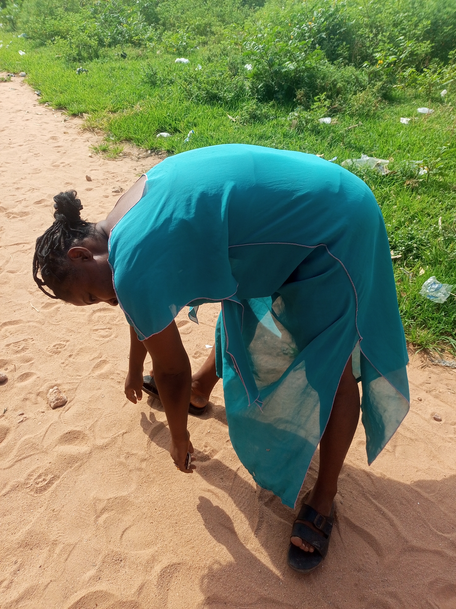 An overhead shot shows a person bent over on a sandy path. They are wearing a teal dress and black sandals. The person's hair is pulled up into a bun. The path is next to a grassy area with some scattered white trash. The lighting suggests it is daytime, and the sun is casting shadows on the sand.