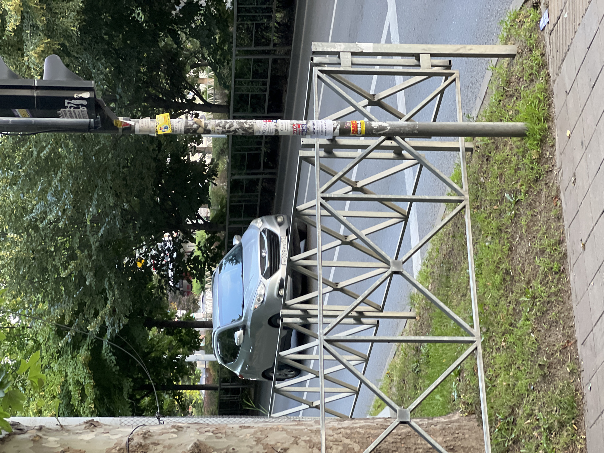 In Krasnodar, Russia, on a paved street, a silver Hyundai Tucson is seen driving on the road. The car has a license plate that reads "W 009 PK 93". In the foreground, a metal fence with a geometric pattern stands on a grassy median. A traffic light pole is visible to the right of the fence, covered in various stickers. Lush green trees line the background, suggesting a city park or tree-lined avenue. The day appears overcast.