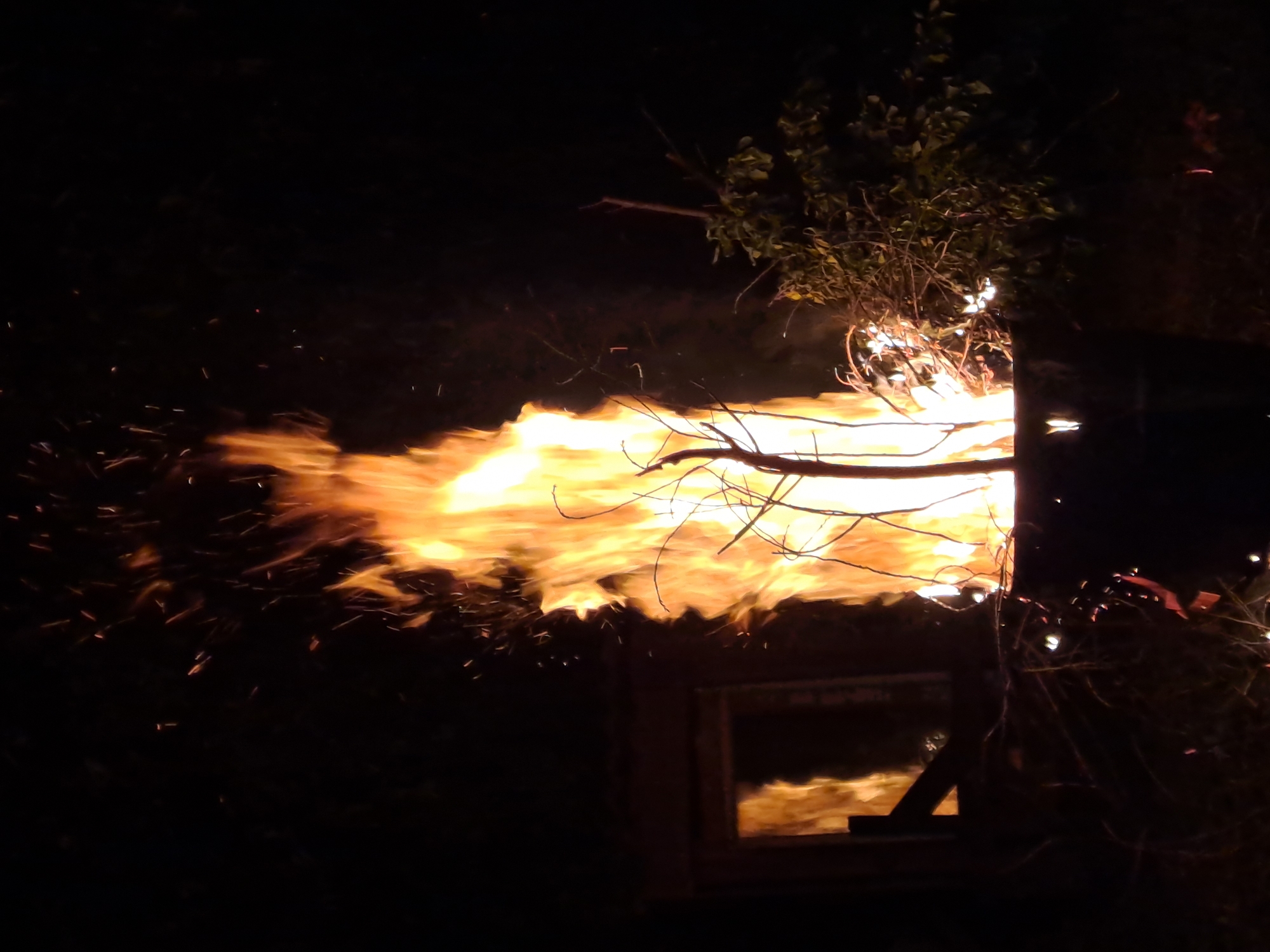 This media file captures a vivid scene of a large outdoor fire burning at night in Полянки, Russia. The primary subject is a substantial fire with bright orange and yellow flames that spread prominently across the lower and central parts of the image, moving from right to left. Numerous glowing embers and sparks are visible, scattering upwards and to the left against the dark background, indicating intense heat and active combustion.
Dark, silhouetted branches and some leafy foliage are visible in the upper right and central portions of the frame, with some branches directly within or extending into the flames. To the far right, a dark, rectangular structure or container is present, possibly a burning shed, a large bin, or a custom-built incinerator. This structure features a distinct, window-like opening near its base, reflecting the intense orange light of the fire.
The setting is clearly outdoors at night, as evidenced by the deep blackness surrounding the illuminated areas of the fire. The activity depicted is a large-scale burning event, possibly a controlled bonfire or the disposal of brush/waste. No people or discernible text are visible in the image. The overall impression is one of raw energy and warmth emanating from the powerful blaze.