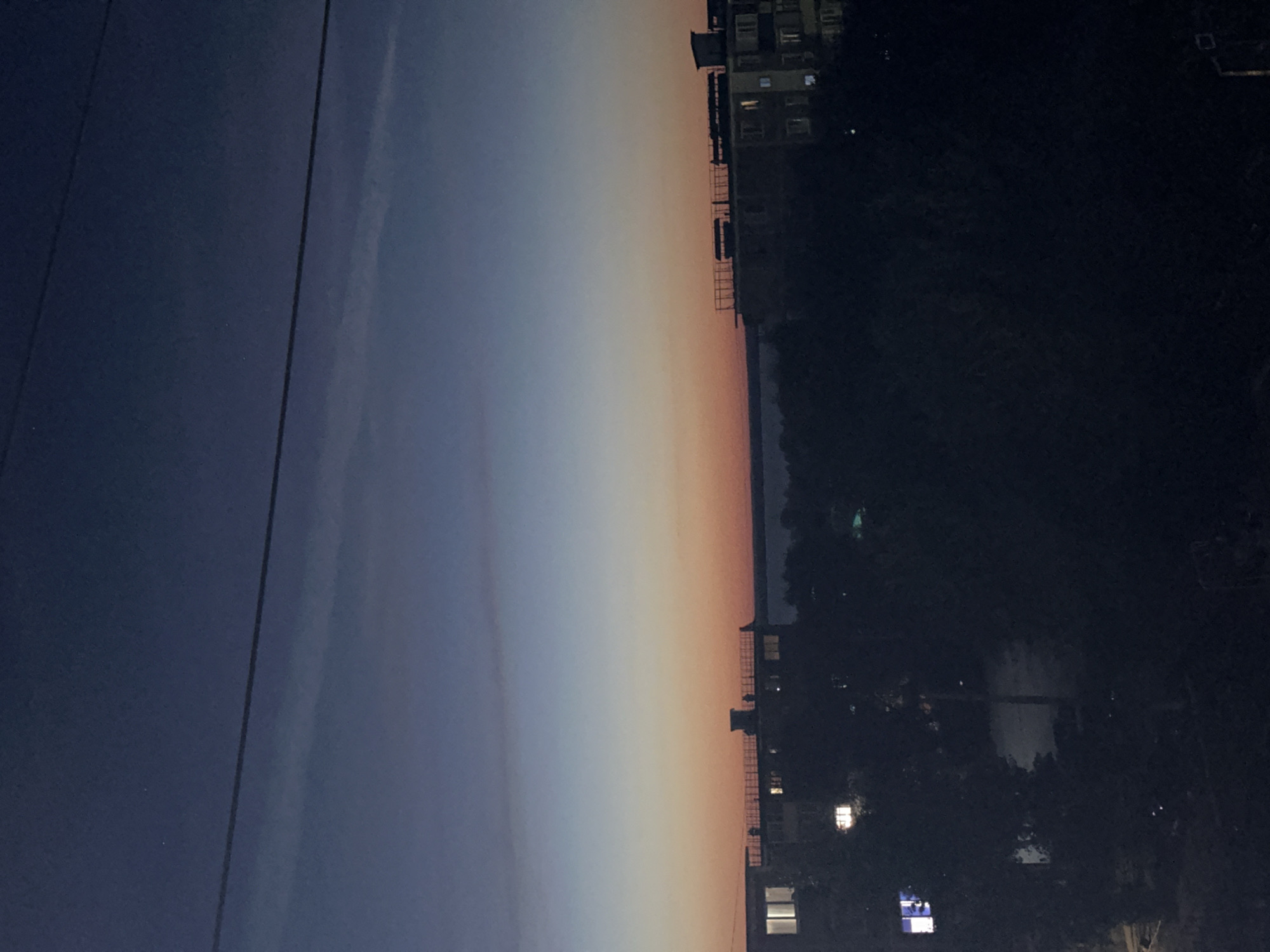 The image captures a twilight scene over the city of Energetik, Russia. The sky transitions from a deep blue at the top to shades of orange and pink near the horizon, suggesting the sun has recently set or is about to rise. A thin, dark horizontal line, likely a power line, stretches across the upper portion of the frame. Below the sky, the dark silhouettes of apartment buildings and trees are visible. Some windows in the buildings are illuminated, indicating activity within. The overall mood is serene and quiet, typical of the transition between day and night in an urban environment.