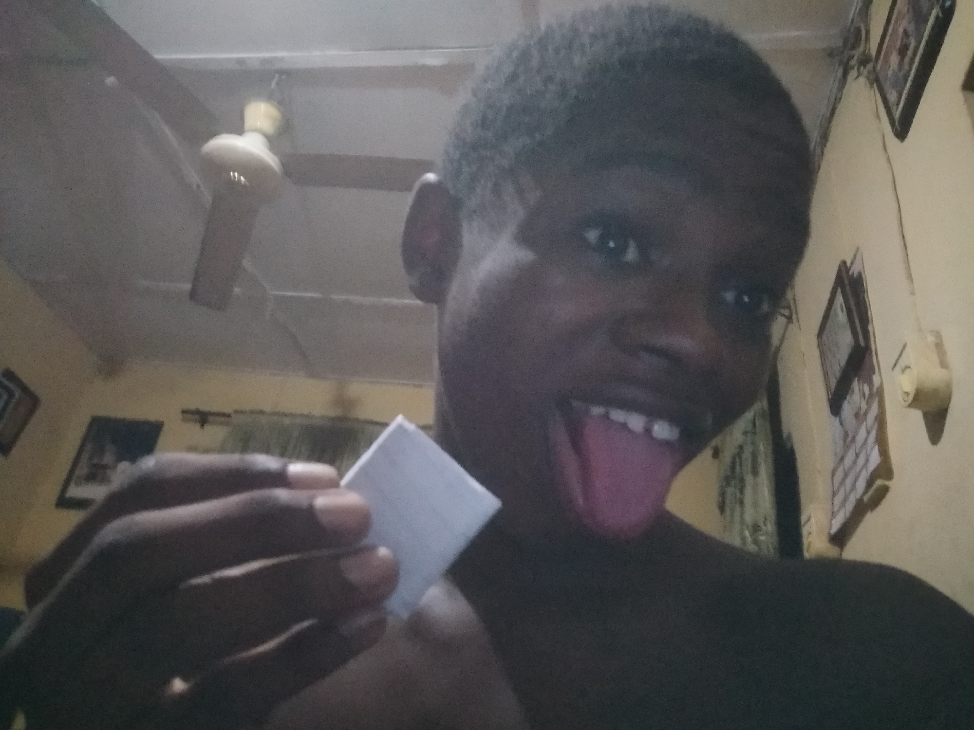 A close-up shot features a dark-skinned person with closely cropped hair, sticking their tongue out. They are holding a small, white rectangular object in their left hand. The background shows an interior space with a ceiling fan, pictures on the wall, and a calendar. The location is Unknown, Unknown.