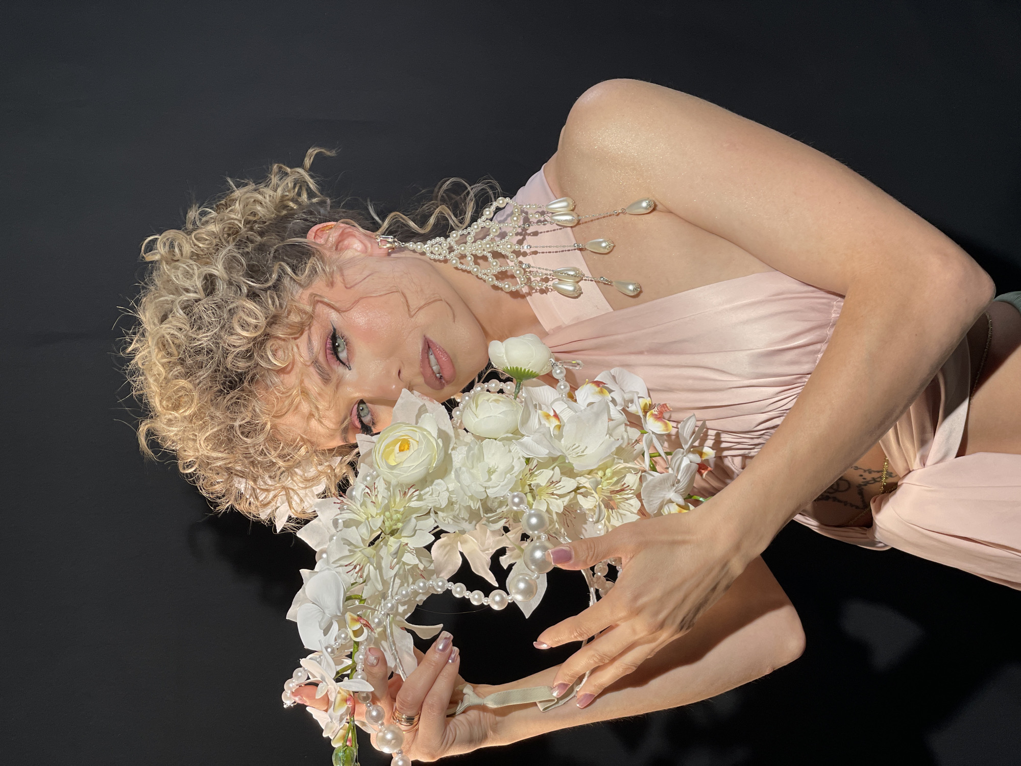 A woman with curly blonde hair is lying on a black surface, holding a floral arrangement of white flowers and pearls. She is wearing a light pink dress and a long, ornate pearl earring. Her makeup includes pink eyeshadow and lip gloss. The image appears to be taken in a studio setting with a dark background. The scene is not located in the city of Дачное, Russia.