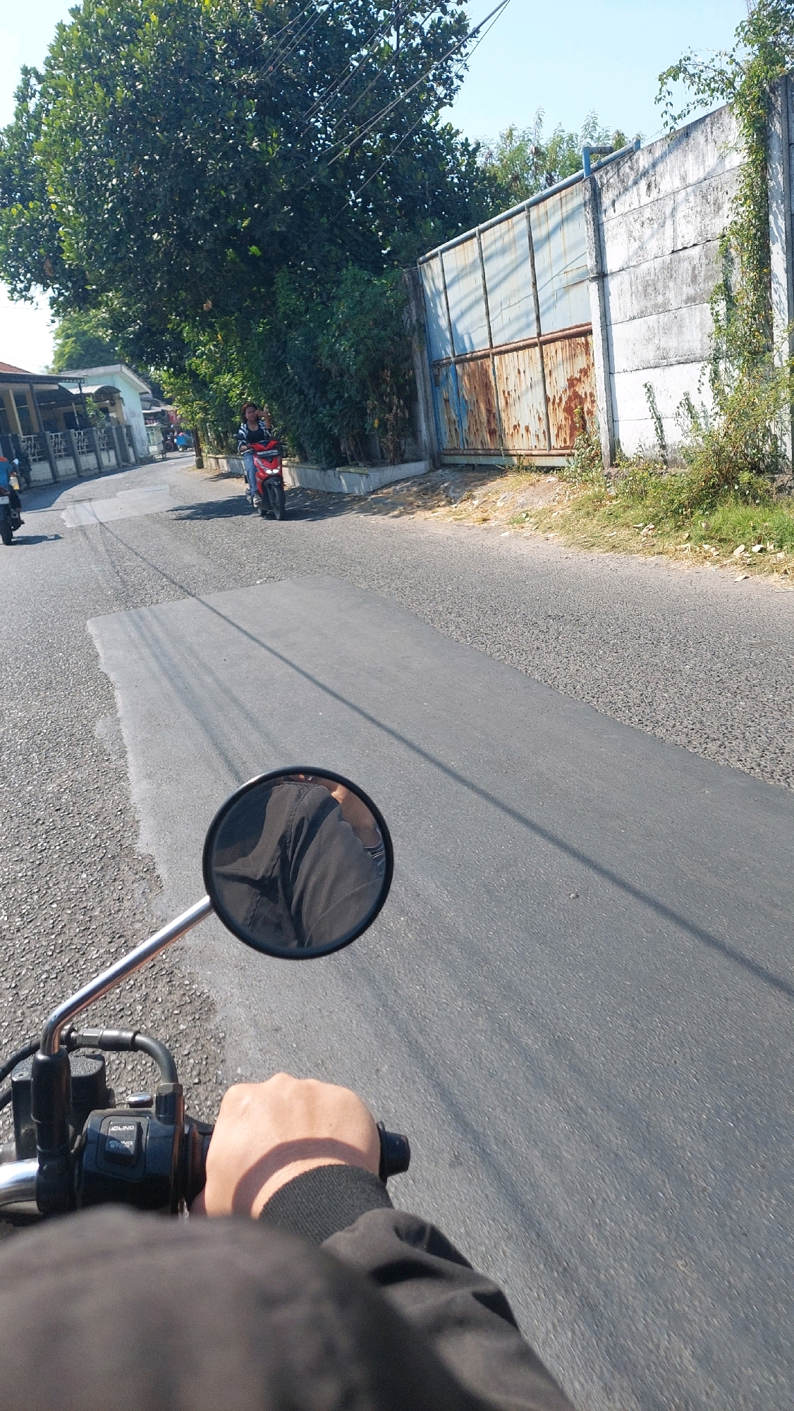 A first-person perspective shot taken from a motorcycle in Sidoarjo, Indonesia. The road is paved with asphalt and has visible lane markings. A large tree with dense foliage is on the left side of the image, with a building and other trees behind it. To the right, there is a tall, weathered concrete wall with a large, rusty metal gate. The gate is partially open, revealing a lighter colored surface behind it. In the middle of the road, a person on a red and black motorcycle is riding away from the camera, with a passenger behind them. A motorcycle rearview mirror is visible in the foreground, reflecting dark clothing. The sky is clear and blue.