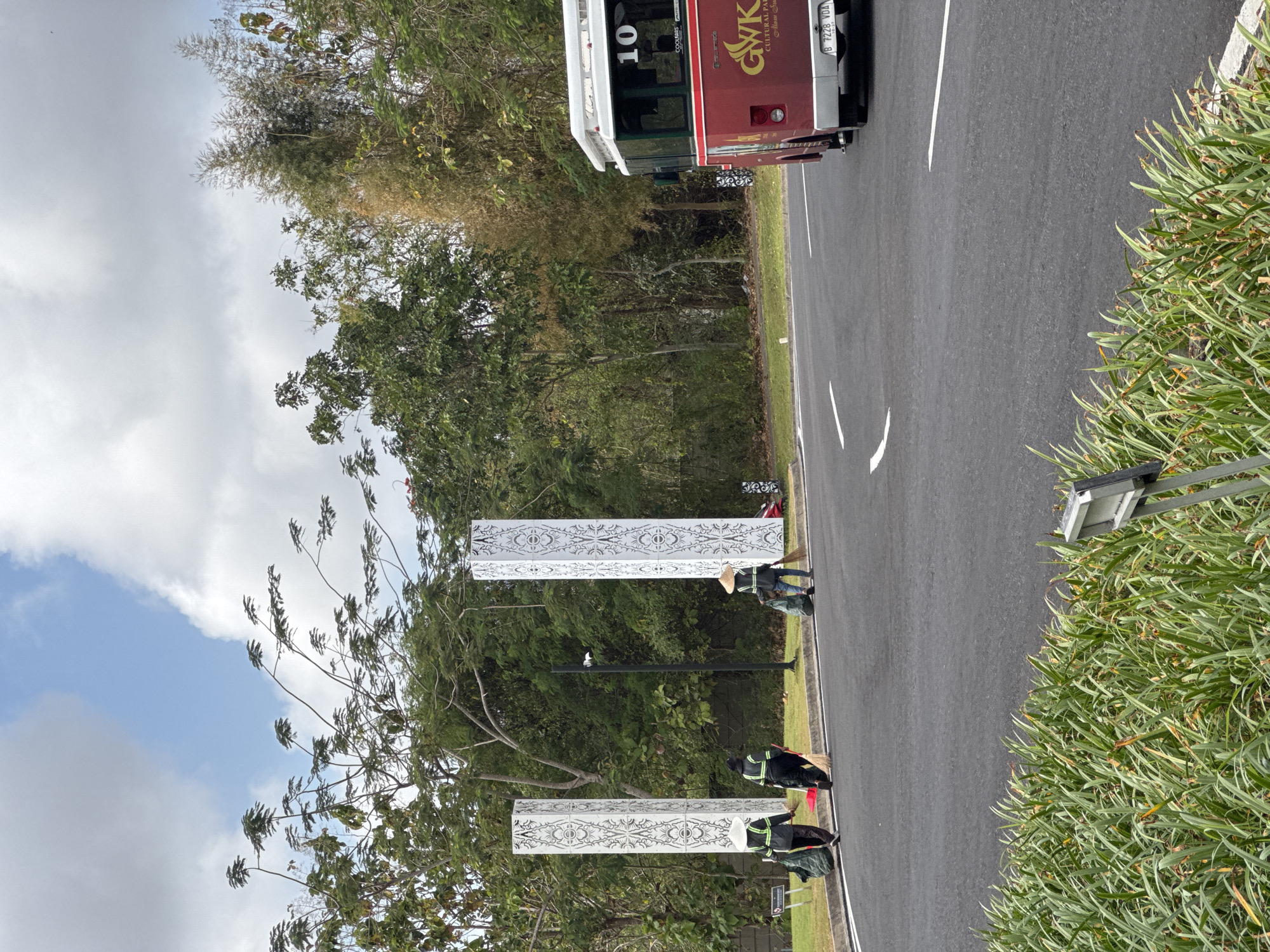 This image depicts the entrance to the GWK Cultural Park in Kuta Selatan, Indonesia. Three individuals, two women and one man, are seen sweeping and collecting debris along the roadside. They are dressed in work attire, with two wearing conical hats. Two tall, white rectangular pillars with intricate patterns flank the roadway, suggesting a formal entrance. A red bus with "10" visible on its side is parked on the right side of the road, with "GWK CULTURAL PARK ALAM SUARA" and a license plate visible. The environment is lush with trees and vegetation surrounding the paved road. The sky is partly cloudy.