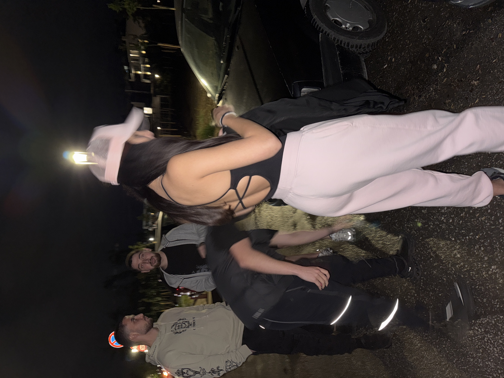 The image depicts a group of four individuals in an outdoor setting at night. A female in a black bodysuit and light pink sweatpants stands closest to the camera. She wears a pink baseball cap. Behind her and to the left are two males. The first is wearing a beige hoodie with graphics and the second is wearing a grey and black jacket. Another male is partially visible between them. The setting appears to be near a street, with a car visible partially in the upper right corner. A blue and white street sign is visible on the bottom left. The ground is paved. Without additional information, the location within Plovdiv, Bulgaria cannot be determined.