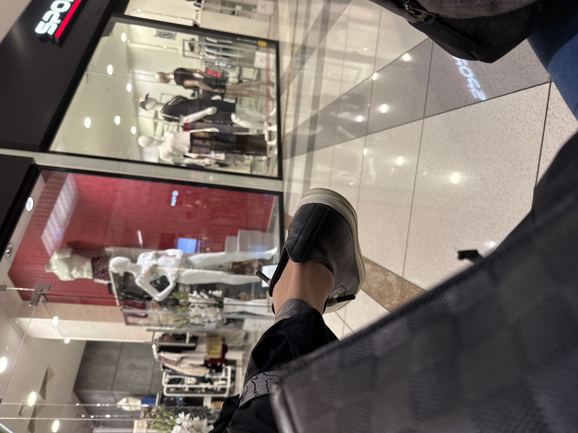 This slightly rotated, low-angle photograph captures a person's foot and a view of a retail store within what appears to be a modern shopping mall in Varna, Bulgaria. The foreground is dominated by the person's left foot, wearing a black slip-on shoe with a white sole, and black trousers. A glimpse of an ankle or lower leg with a dark pattern, possibly a tattoo or strap, is visible. Part of a dark, checkered bag or garment is seen in the bottom right corner.
In the background, a brightly lit clothing store is visible through its expansive glass window. Several mannequins, dressed in various outfits, are arranged within the display, showcasing items like shirts, dresses, and trousers. The store's interior features white walls, clothing racks, and embedded spotlights. Above the window, a black sign with illuminated red and white letters partially reads "SPOR", likely part of a brand name such as "SPORT". Reflections of overhead lights and faint, inverted text, possibly reading "POSE" or "ROSE" from the sign, are visible on the polished, light-colored tiled floor. The floor has contrasting darker gray sections, contributing to the modern aesthetic of the mall environment. The bright, consistent lighting suggests an indoor setting during operational hours.