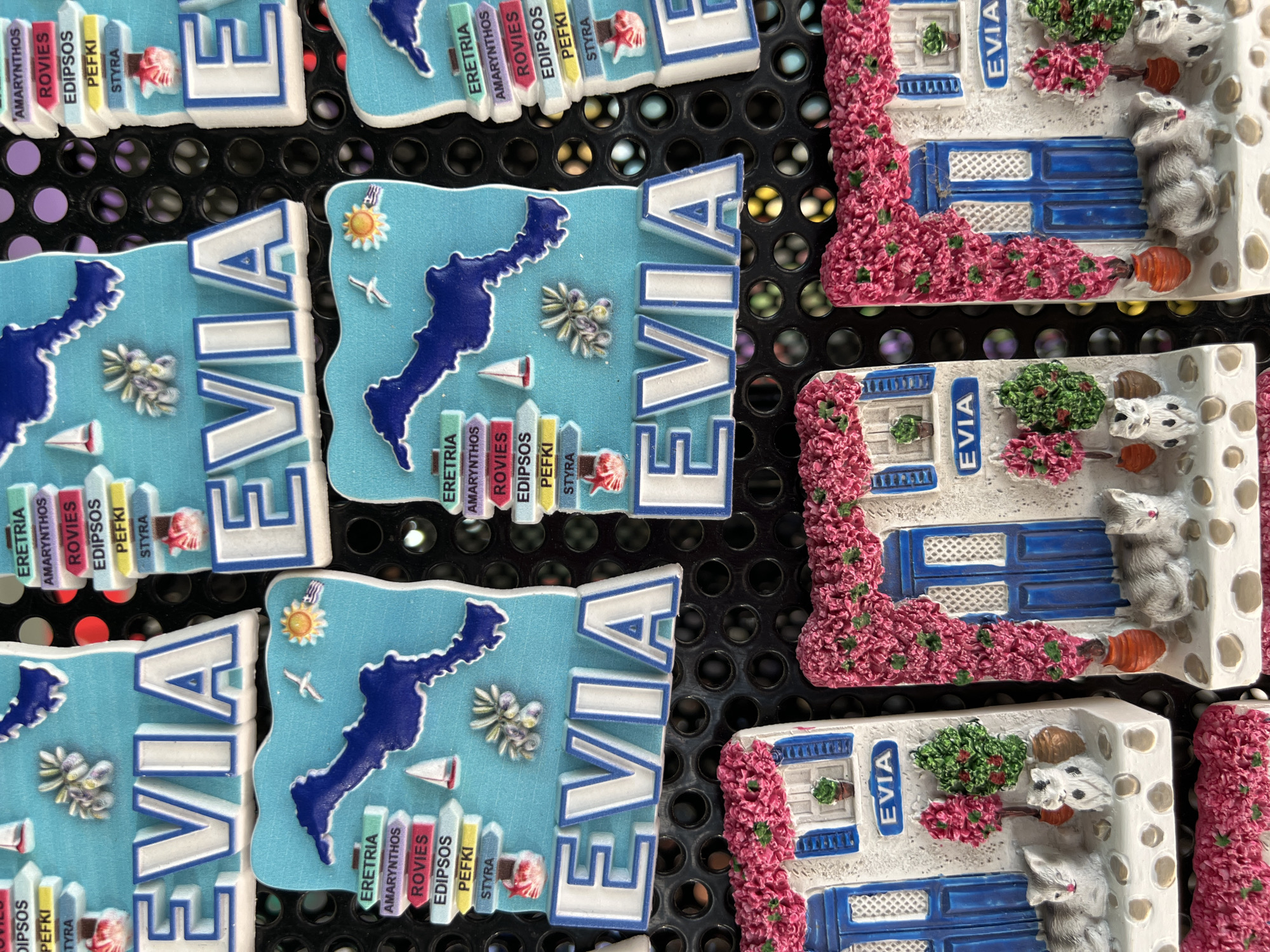 The image displays a collection of souvenirs, likely refrigerator magnets, arranged in rows. The dominant design features a light blue background shaped like the island of Evia, with the word "EVIA" prominently displayed in raised white lettering with blue outlines. Accompanying the island shape are graphics of sailboats, a sun with the Greek flag, and clusters of purple olives. Stacked road signs point to various locations, including "ERETRIA," "AMARYNTHOS," "ROVIES," "EDIPSOS," "PEFKI," and "STYRA."
Below these island-shaped magnets are smaller souvenir designs depicting white houses with blue doors and windows, adorned with vibrant pink bougainvillea. Small potted plants and two ceramic cats are placed on the doorstep of each house. The name "EVIA" is also visible on the facade of these houses. The souvenirs are placed on a black, perforated surface, suggesting a display rack in a shop. The overall impression is a retail display of tourist memorabilia.