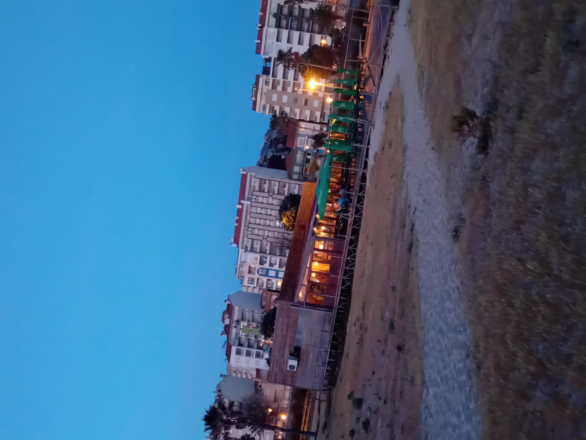 The image depicts a dusk scene in Figueira da Foz, Portugal. The view includes residential buildings, a beachside restaurant with visible interior lighting, green sun umbrellas, and a sandy path alongside a grassy area. The sky is a gradient of blue, indicating twilight. There are no visible people or specific actions taking place in the immediate frame. Palm trees and a street lamp are present near the buildings.