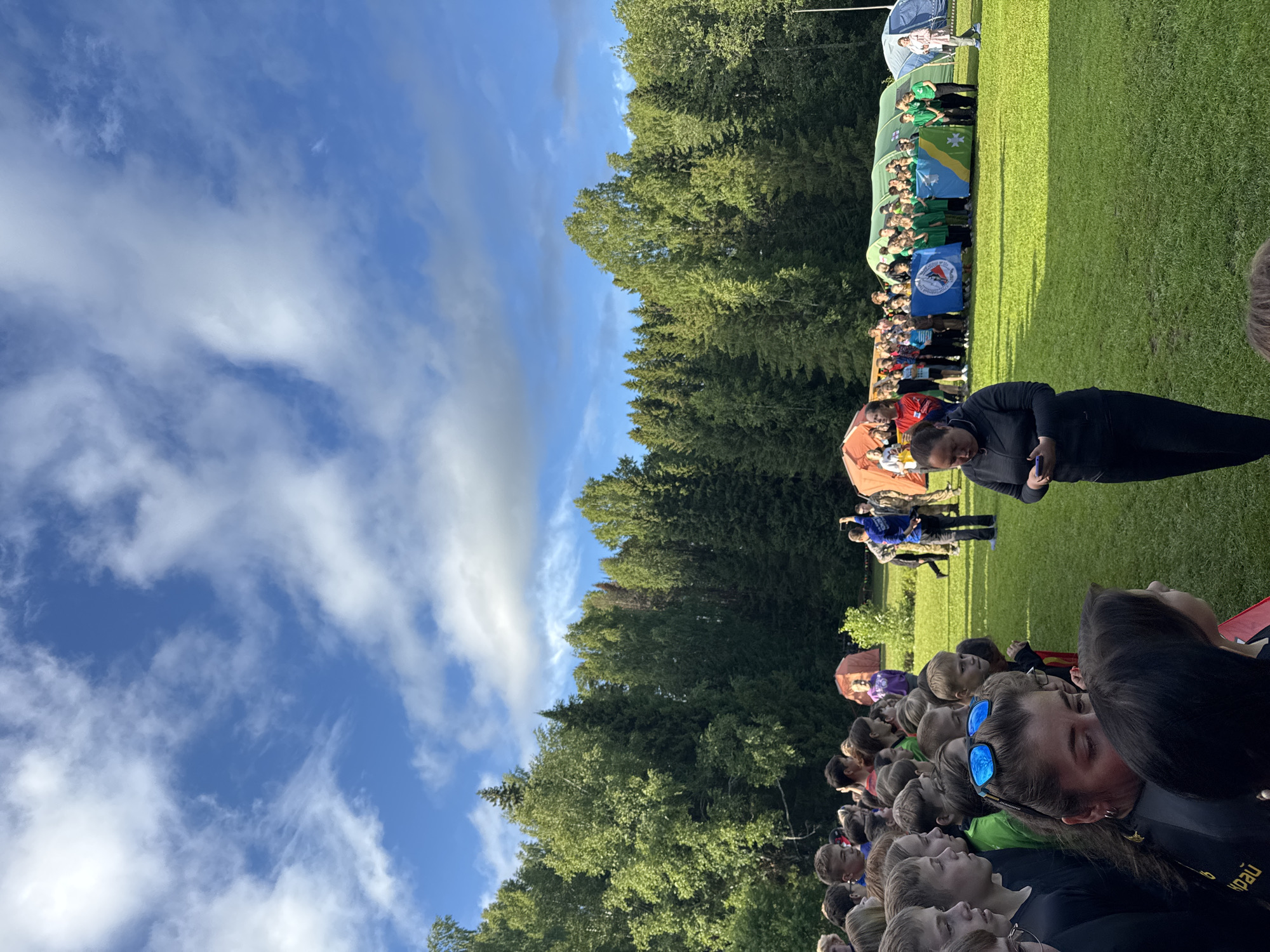 This media file depicts an outdoor gathering of people in a grassy field, with a dense forest bordering one side and a bright blue sky with scattered clouds above. The scene appears to be set in Sergino, Russia, suggested by the text on one of the flags.
The subjects are predominantly a large group of people, many of whom appear to be children and adolescents, standing in rows. They are facing towards the forest and the other side of the field. Some individuals are wearing matching green shirts, suggesting a team or group affiliation. There are also tents or canopies set up, one of which is blue and white, and another appears to be orange. Several flags are displayed, including a blue circular flag with white text and an emblem, and another flag with a green and yellow design featuring a star-like symbol.
The activity taking place seems to be some form of outdoor event or ceremony, possibly a competition or festival, given the organized rows of people and the displayed flags. The individuals in the foreground are looking upwards and towards the right of the frame, their expressions generally neutral or expectant. The sun is visible, casting shadows on the grass, indicating it is daytime. The weather appears to be pleasant and sunny.
Notable details include the clear blue sky with wispy white clouds, the vibrant green of the grass and the dense foliage of the coniferous trees. The presence of flags and organized groups suggests a communal event. The time of day is likely mid-morning to late afternoon, judging by the angle of the sun and the bright lighting.
Visible text includes Cyrillic script on a black garment in the foreground. One of the flags also has visible text on its blue circular emblem, though it is difficult to read clearly.