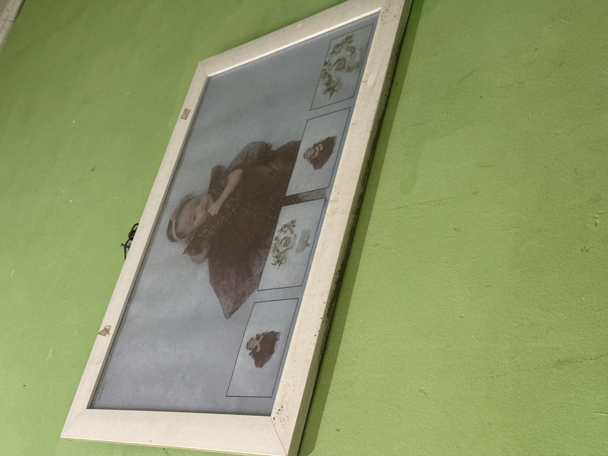 An unframed photograph of a young child is displayed on a light green textured wall. The photograph features a central image of the child wearing a dark red garment and a headband. To the bottom of the central image, there are three smaller inset images, each with a small portrait of a child within a decorative border. The photograph is hung with a black wire. The wall shows evidence of age and wear, with some scuff marks and slight discoloration. The lighting in the image is somewhat dim, casting subtle shadows. There is no indication of the specific location within Babahjurong, Indonesia.