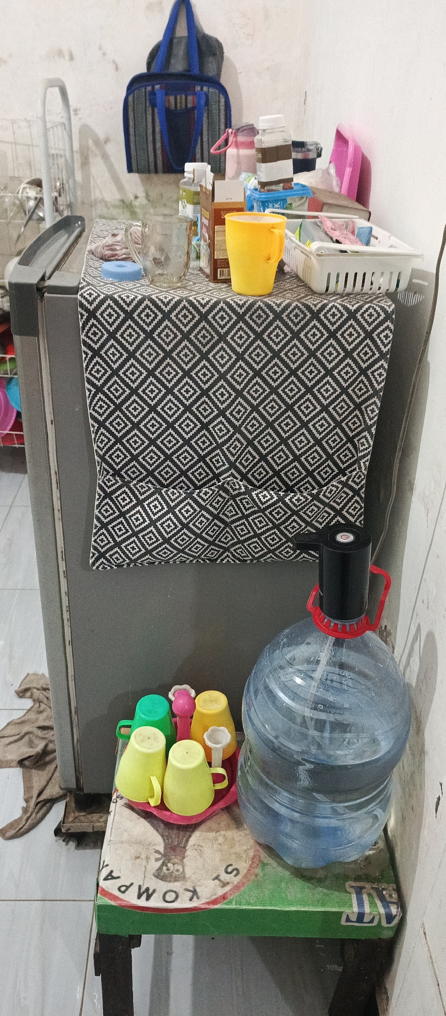 The image captures a still life scene on top of a gray refrigerator in what appears to be a residential setting in Arjasa, Indonesia. The refrigerator is covered with a black and white patterned cloth. On the cloth, various items are arranged: a clear glass mug, a brown box, a yellow cup, and a white basket containing what seem to be small packets. Behind these items, a blue and gray duffel bag is hanging on a wall. To the right of the refrigerator, a large, clear plastic water jug with a black pump dispenser is prominently displayed. In front of the water jug, a tray holds several colorful plastic cups in shades of green, yellow, and pink. The overall impression is one of everyday domesticity, with items for immediate use placed on an accessible surface. The lighting suggests it is daytime, with light entering from an unseen source.