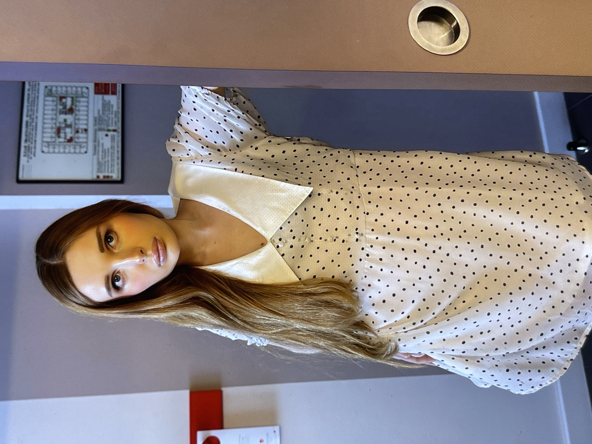 A medium shot shows a young woman with long, light brown hair and fair skin, wearing a white polka-dot dress with a large collar. She stands in a doorway, leaning against the frame. Her face is turned towards the camera, and she is looking directly into the lens with a neutral expression. Her makeup includes rosy cheeks and light pink lipstick. To her left, a portion of a white wall with a red and white sign is visible. Behind her, a muted blue wall displays a framed floor plan. To her right, a brown doorframe with a circular brass handle is present. The overall lighting is soft, with some warm tones highlighting the woman's face.