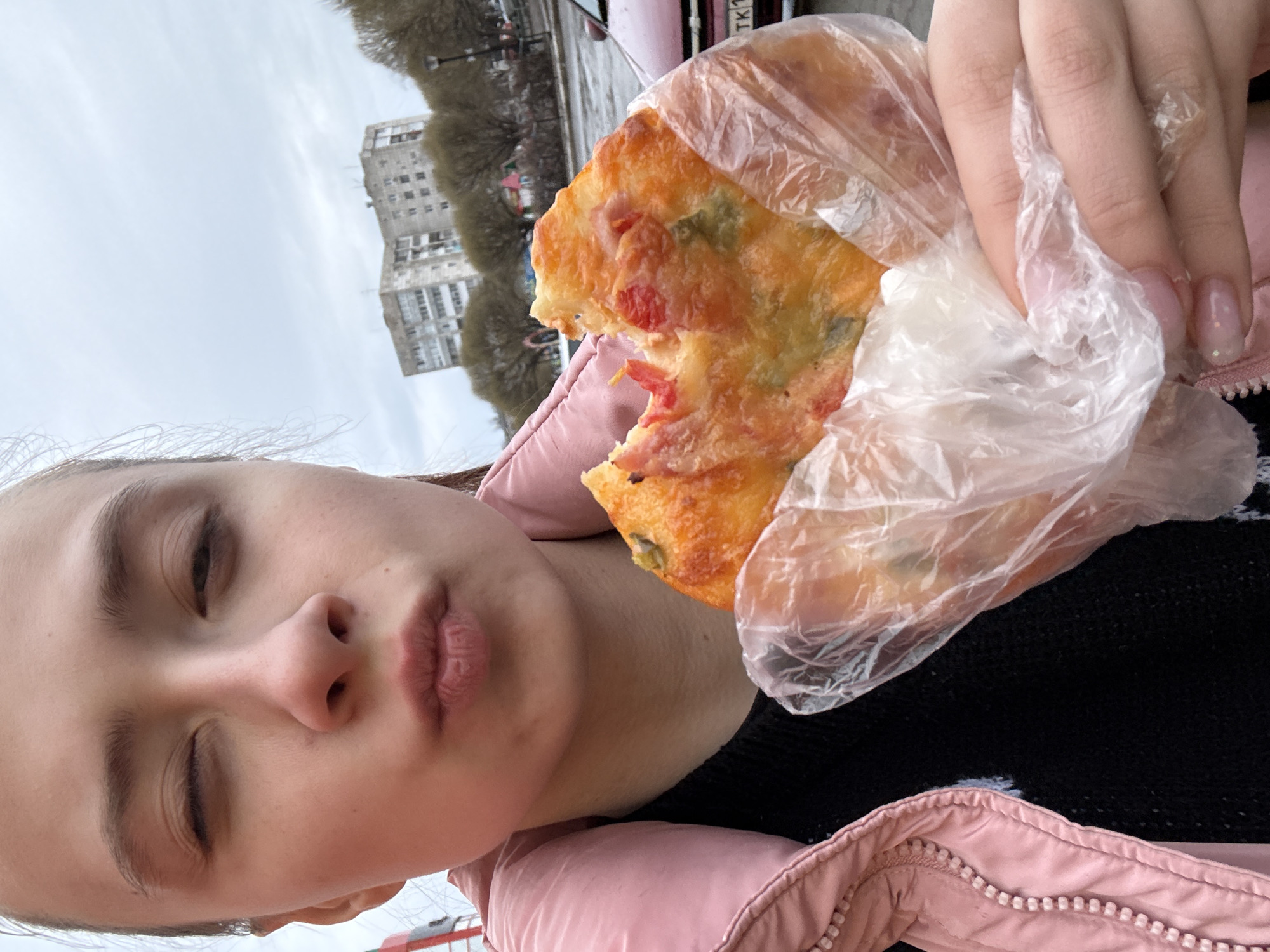 A photograph depicts a person holding a slice of pizza in a plastic bag. The individual has fair skin, visible eyelashes, and a pink jacket. The pizza slice is topped with cheese, tomato, and what appears to be green pepper. In the background, there is a cityscape, trees, and a pink-colored vehicle. Based on the presence of buildings and trees, the setting appears to be urban. The photograph may have been taken in Chaikovsky, Russia.