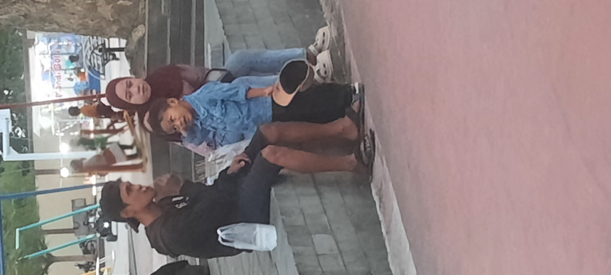 In Kanigoro, Indonesia, a young boy in a blue denim shirt and black pants stands holding a dark-colored hat, looking to his left. To his left, a young man wearing a black hoodie and dark shorts sits on a concrete ledge, his right hand in his pocket, and his gaze directed away from the camera. A woman wearing a maroon hijab sits behind the boy, holding him, and is looking forward. A white plastic bag with items inside is placed on the concrete ledge to the left of the seated man. In the background, a playground structure with swings and a ride is visible, along with trees and what appears to be a vendor stall or building. The ground in the foreground is a textured pink surface.