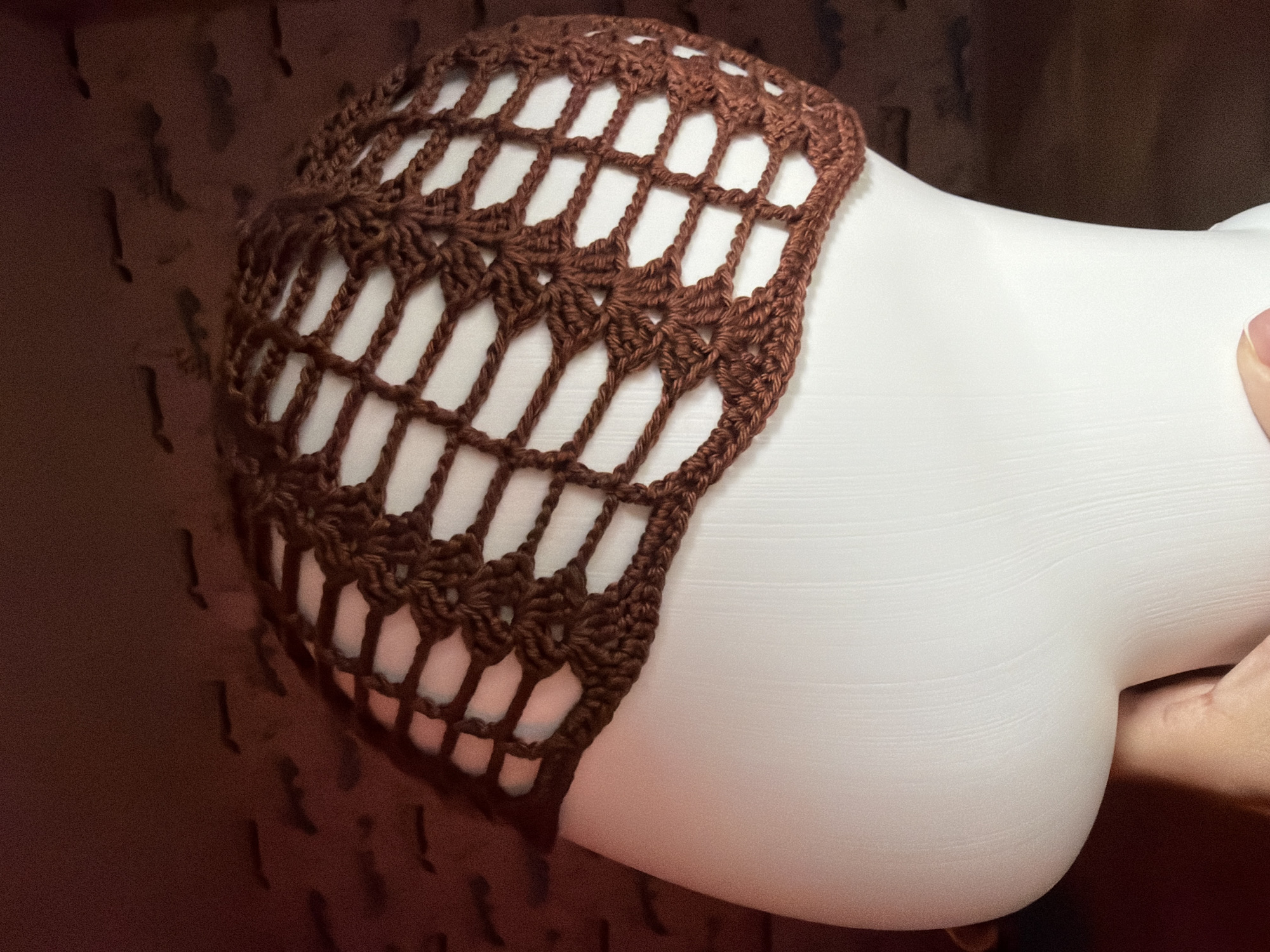 A close-up studio shot shows a brown crocheted net or cap stretched over a white, stylized mannequin head. The mannequin head is being held by a hand, seen on the right side of the image. The background consists of blurred brown tones suggesting a textured wall or surface. The crocheted item features a repeating pattern of interconnected geometric shapes, creating a grid-like structure. The lighting is soft, accentuating the texture of the crochet work and the smooth surface of the mannequin head. There is no indication of this image having been taken in Chaikovsky, Russia.