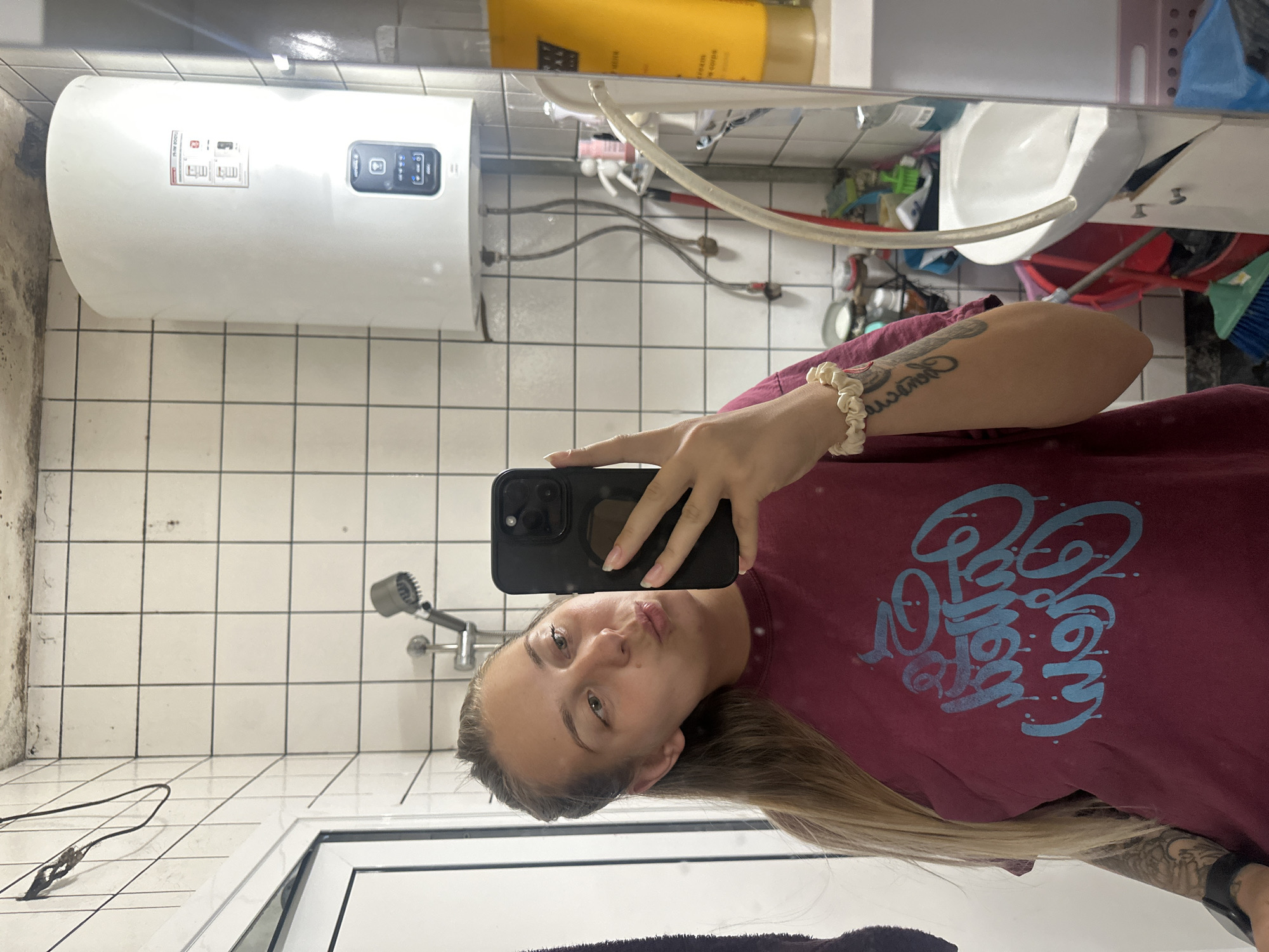 A young woman with blonde hair pulled back and wearing a maroon t-shirt with blue graffiti-style lettering stands in a bathroom and takes a selfie. She is puckering her lips, and her left arm is extended, holding a black iPhone. She has a scrunchie on her right wrist and tattoos on her left arm. The bathroom appears to be in a state of disrepair, with visible mold on the ceiling and water stains on the walls. A white water heater is mounted on the wall, and a showerhead is visible. In the background, a white sink is attached to a cabinet, and various cleaning supplies and toiletries are scattered around. The overall scene suggests a utilitarian and possibly low-income living situation in Plovdiv, Bulgaria.