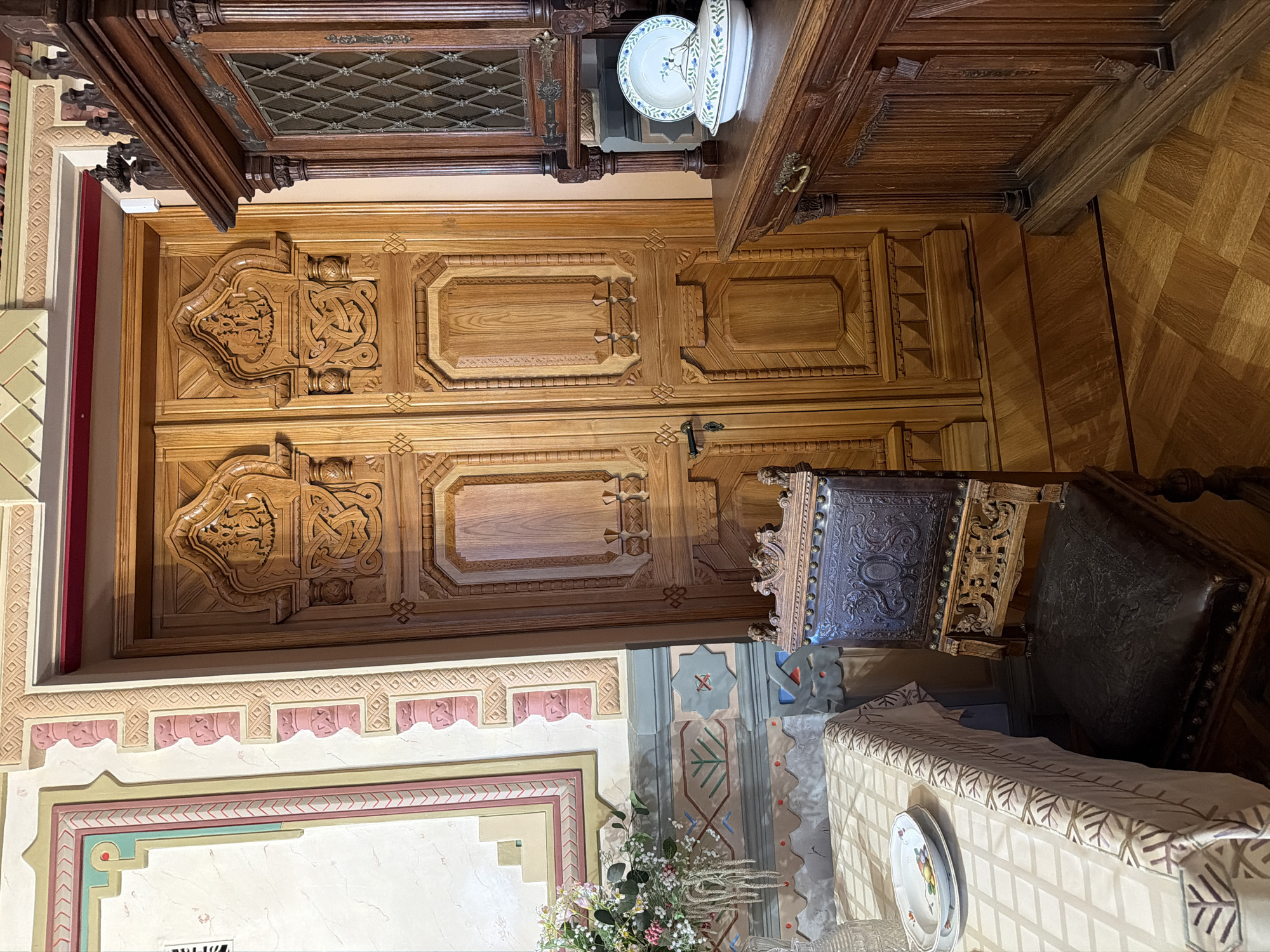 The image depicts an interior scene featuring ornate wooden furniture and decorative walls. In the foreground, a table is partially visible, covered with a patterned tablecloth, and set with two plates. To the right of the table, a dark leather-upholstered chair with intricately carved wooden elements stands prominently. Behind the chair is a tall, elaborately carved wooden double door. To the right of the door, a substantial wooden cabinet or sideboard is present, with a glass-paneled section and a decorative top. A plate and a lidded serving dish with blue floral patterns rest on the surface of the cabinet. The walls are adorned with decorative molding and painted patterns, featuring geometric shapes and floral motifs. The floor is covered in parquet flooring. The scene appears to be staged in a room with historical or artistic significance. While the image itself does not contain explicit indicators of its location in Nizhny Novgorod, Russia, it is consistent with the interior of a museum or historic building in the city, such as the State Museum-Reserve "Kremlev-Nizhny Novgorod" or a similar heritage site known for its preserved interiors and decorative arts.