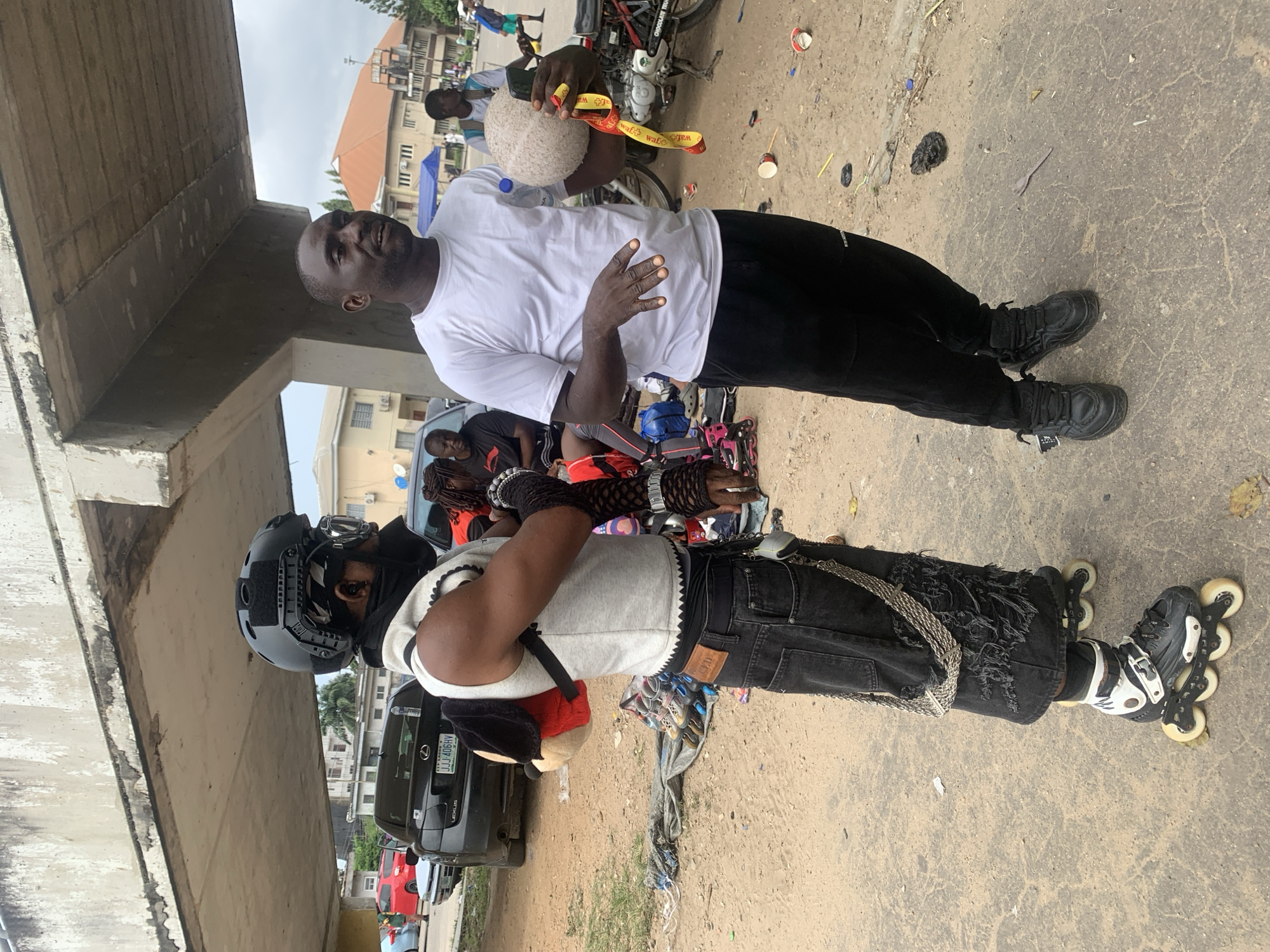 Two men are standing on a dirt and concrete surface near a highway overpass in Alagbede, Nigeria. The man on the left is wearing a black helmet, goggles, a grey vest over a white shirt, black ripped jeans with a prominent silver chain, and rollerblades. He has a plush toy attached to his backpack. The man on the right is wearing a white t-shirt and black pants. He is holding a round, textured object and a yellow and red ribbon. In the background, there are several vehicles, including a black Lexus sedan, and various buildings. The ground is littered with debris, including plastic cups and wrappers. The sky is overcast.