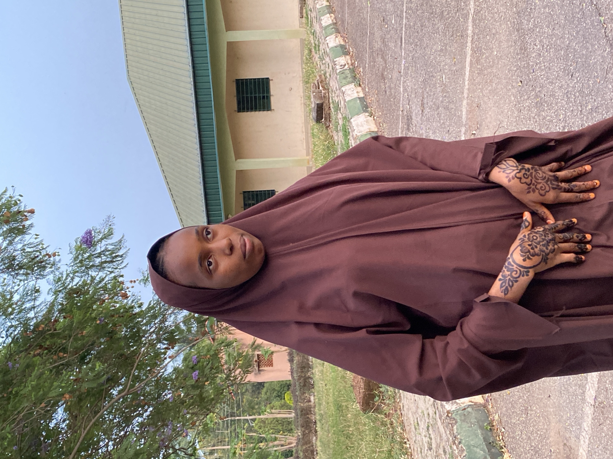 A young woman wearing a dark brown hijab stands outside. She is holding both hands up, displaying henna tattoos on the back of each hand and orange paint on her fingernails.
The background shows a building of light tan color with a horizontal green stripe, possibly a government building, and a sidewalk. Green vegetation and trees are also visible. The location is Vom, Nigeria.