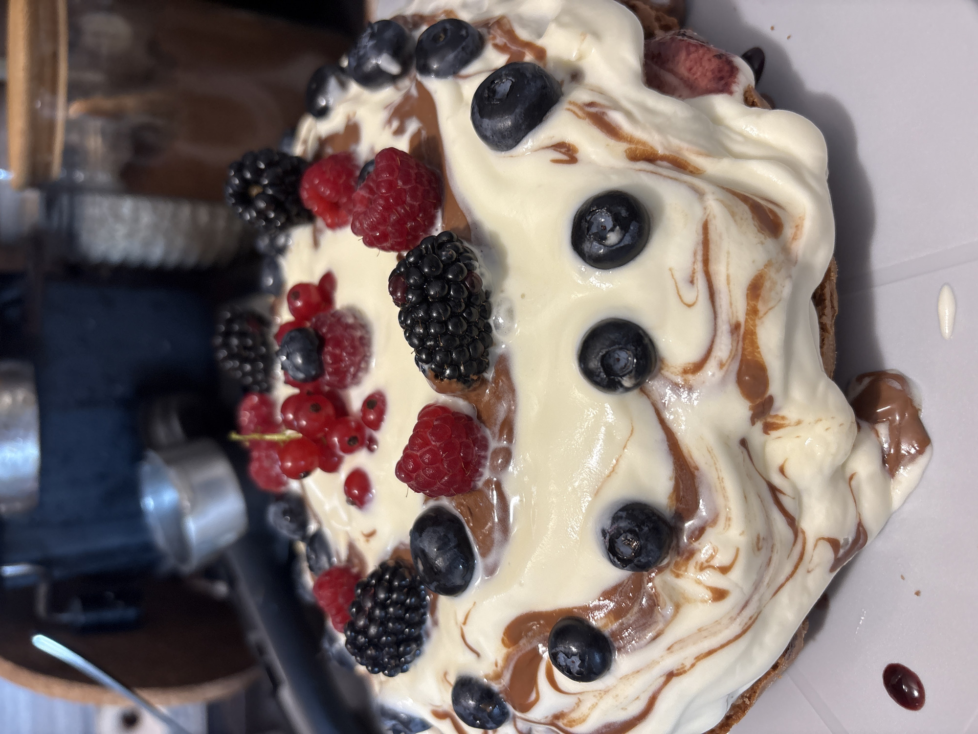 A close-up shot captures a dessert topped with whipped cream and various berries, including raspberries, blackberries, blueberries, and red currants. The dessert is artfully decorated with swirls of chocolate sauce, which also drips down the side, pooling slightly on the white surface below. In the blurred background, indistinct objects suggest a kitchen or dining setting, possibly with kitchen appliances and containers. The lighting is direct and bright, highlighting the textures and colors of the food.