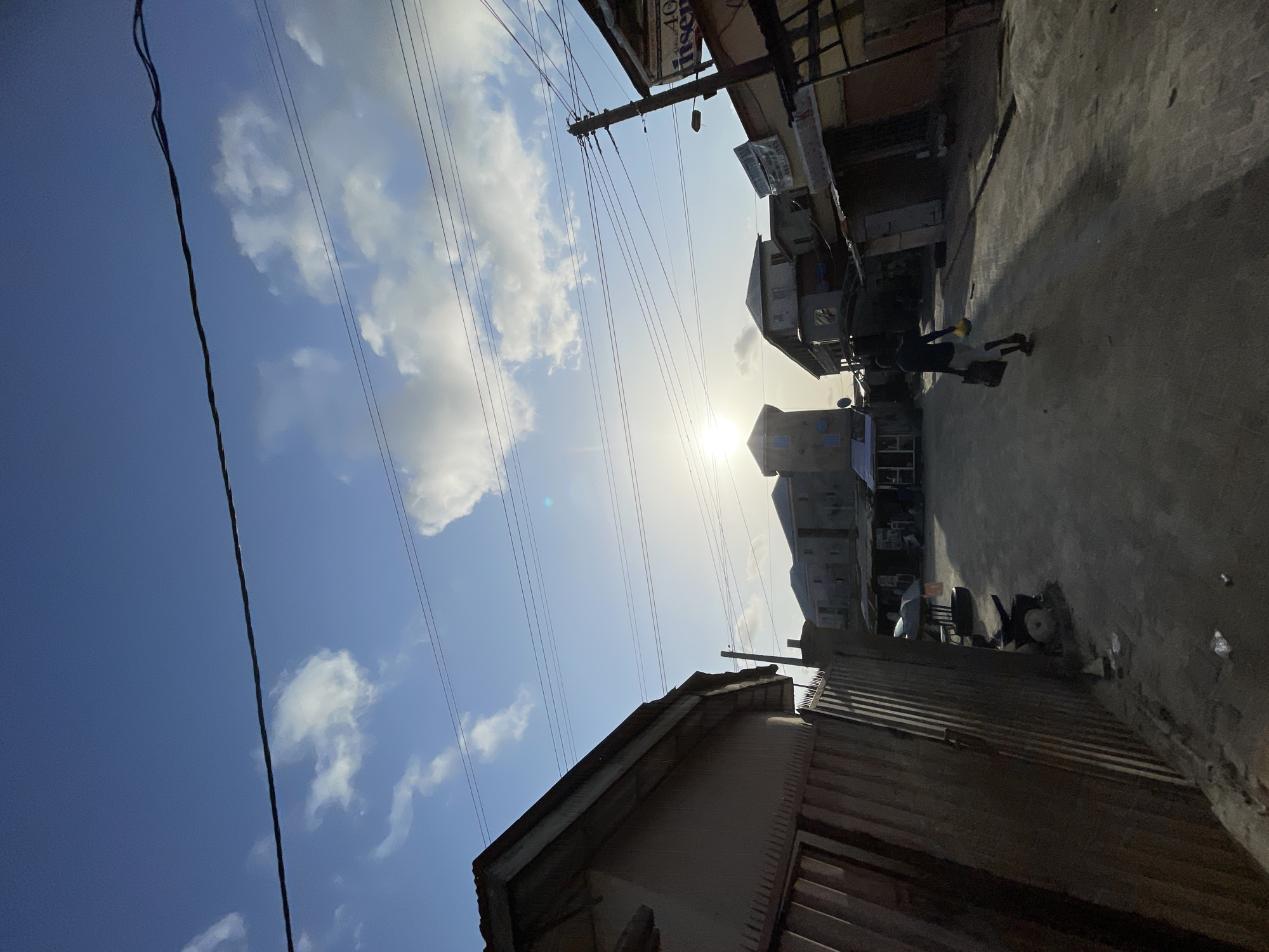 The image depicts a street scene in Ado, Nigeria. The view is tilted, showing a blue sky with scattered clouds and the sun obscured by buildings in the distance. Power lines stretch across the sky. On the right, buildings line the street with signs visible. A person walks along the street. The foreground shows rooftops and structures.