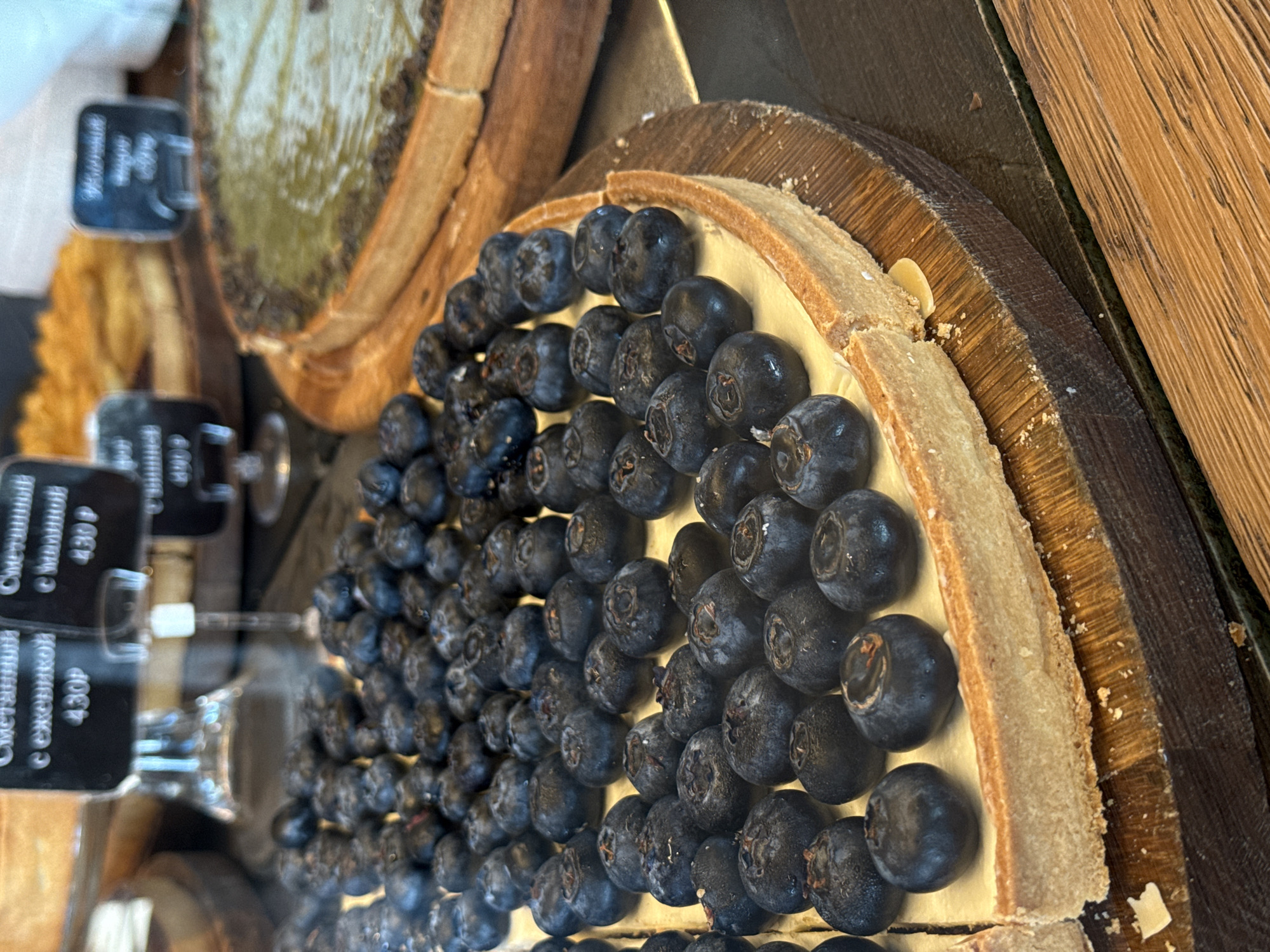 This close-up image captures a display of tarts in what appears to be a bakery or cafe in Sochi, Russia. The primary subject is a large, round tart adorned with numerous fresh, dark blue blueberries covering its surface. The blueberries rest on a light-colored, creamy filling, possibly a custard or sour cream base, encased in a thick, golden-brown pastry crust. The tart is placed on a rustic wooden board, which in turn sits on another textured wooden surface, suggesting a natural or artisanal aesthetic for the display.
To the left and slightly behind the blueberry tart, a portion of another circular tart is visible, featuring a distinct greenish top, possibly indicative of a matcha or pistachio flavor. Adjacent to these baked goods are several black price tags mounted on thin stands.
Visible text on the tags, written in Russian, provides details and pricing. One clear tag reads "Сметанный с ежевикой 430₽" (Sour cream with blackberry 430₽), while another partially visible tag above it appears to state "Сметанный с малиной 430₽" (Sour cream with raspberry 430₽). These labels, while priced at 430 Russian Rubles, likely refer to other items in the display rather than the prominent blueberry tart. No people are visible, and the scene implies a quiet moment at a food establishment, showcasing its offerings. The lighting suggests an indoor setting with good illumination, highlighting the textures and colors of the tarts.