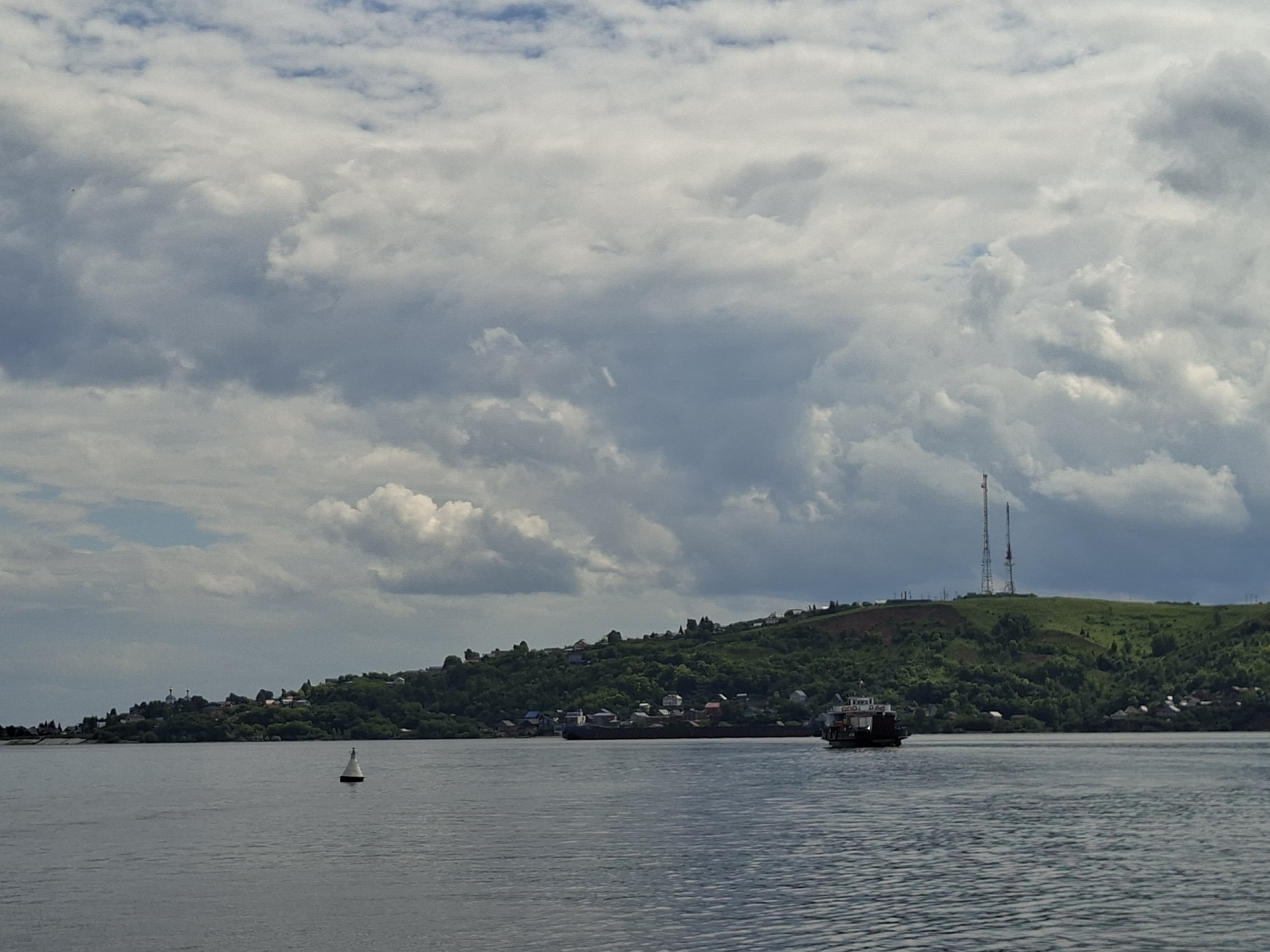 The image captures a wide shot of a calm river on a cloudy day, likely in Kazan, Russia, given the context. The foreground is dominated by the rippling water of the river, reflecting the overcast sky.
In the mid-ground, a white buoy with a black top floats on the water, marking a point of navigation. Further on, a long, dark barge is visible moving along the river. On the right side of the barge, a smaller vessel with a white superstructure is also present.
The background features a lush, green, and hilly landscape that slopes down to the riverbank. A scattering of houses and buildings are visible nestled amongst the trees and on the slopes, suggesting a populated area. Atop the highest hill, two tall communication towers stand prominently against the sky, a clear indication of the location. The clouds are a mix of gray and white, with patches of blue sky peeking through, indicating a dynamic and potentially changeable weather pattern. The overall atmosphere is serene and suggests a peaceful day on the water. There is no visible text in the image.