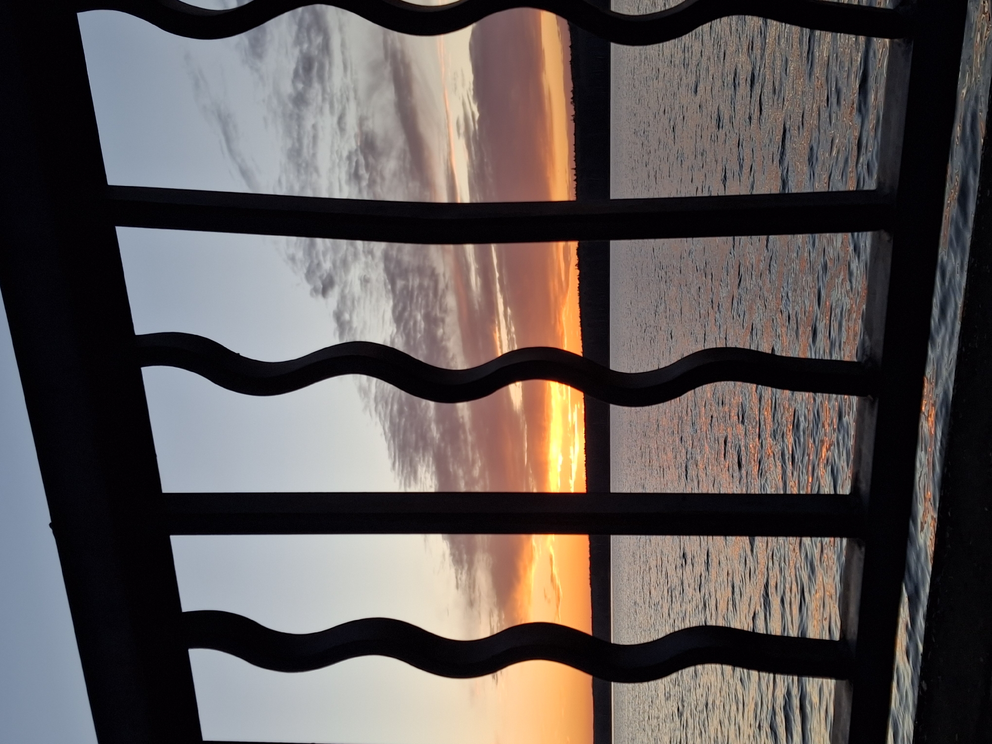 The image shows a view of a body of water (likely the Kama River) at sunset, framed by a dark metal railing with a wave-like design. The sky is a mix of colors, transitioning from a light blue to orange and red hues at the horizon, with some visible clouds. The water reflects the sunset colors. A dark strip of land, possibly forest or shoreline, is visible on the horizon. The railing is in the foreground and dominates the lower half of the image. The location appears to be Chaikovsky, Russia.