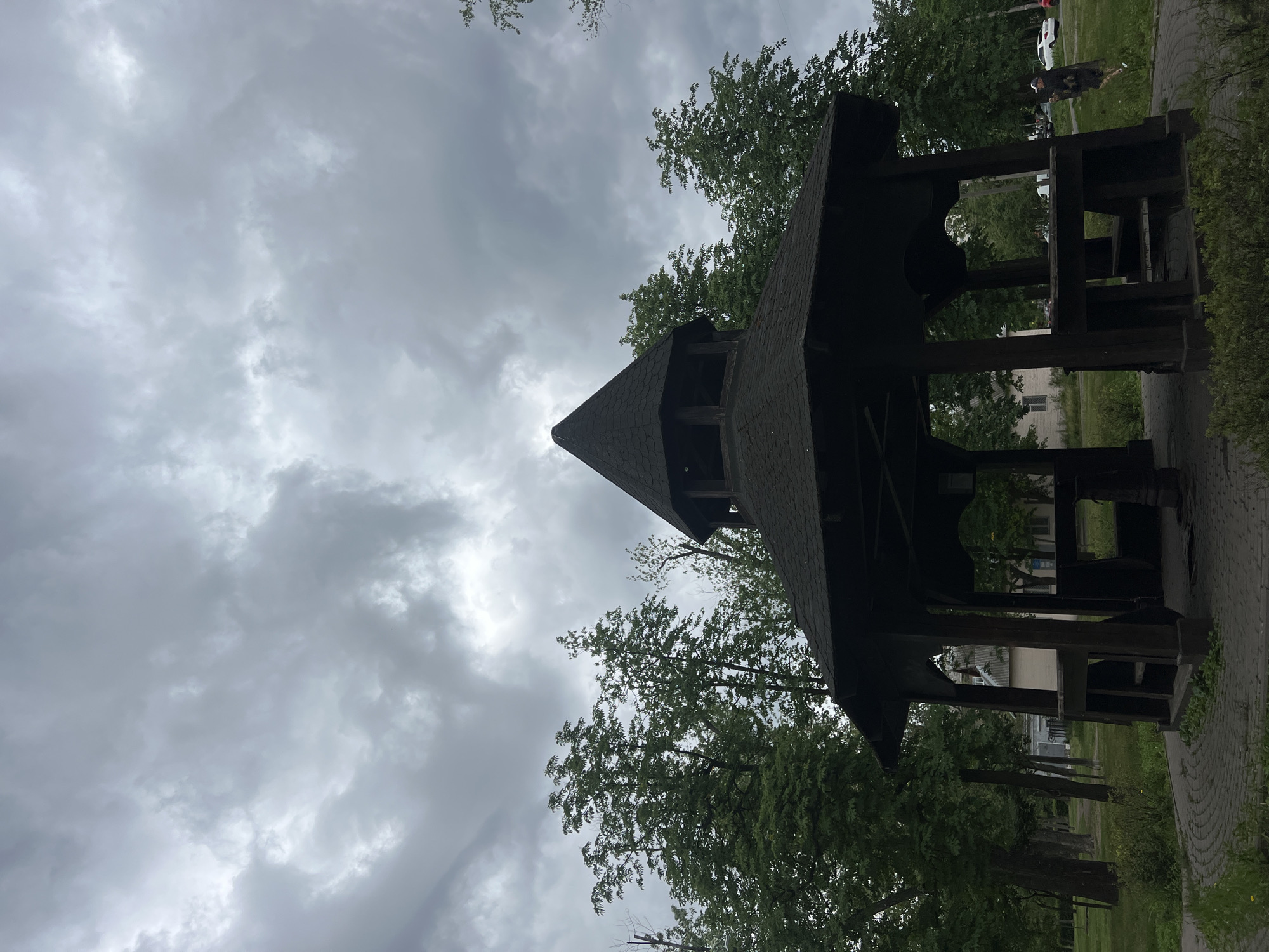 The image, correctly oriented, presents a low-angle view of a dark wooden gazebo or pavilion on a heavily overcast day in Kyiv, Ukraine. The gazebo, appearing on the right side of the frame, features a distinctive multi-faceted, pointed roof with dark shingles and intricate wooden carvings along its eaves and support beams. Much of the structure's interior is cast in shadow due to the dark wood and overhead perspective.
The sky, filling the left and upper portions of the image, is a canvas of dense grey clouds, varying in tone from dark, brooding masses to lighter, brighter patches, suggesting a very cloudy or possibly pre-storm atmosphere. Green foliage from trees is prominent, framing the gazebo and reaching into the sky, with their leaves appearing vibrant against the muted sky.
In the background, visible through the open structure of the gazebo, are hints of distant buildings, possibly residential, and more trees. A paved path or ground made of pavers runs along the lower part of the image, suggesting a park or public area. A faint outline of a person and what appear to be parked cars are visible in the far distance on the right, beyond the gazebo. The overall mood is subdued and naturalistic, emphasizing the architectural detail against the dramatic sky. No text is visible.