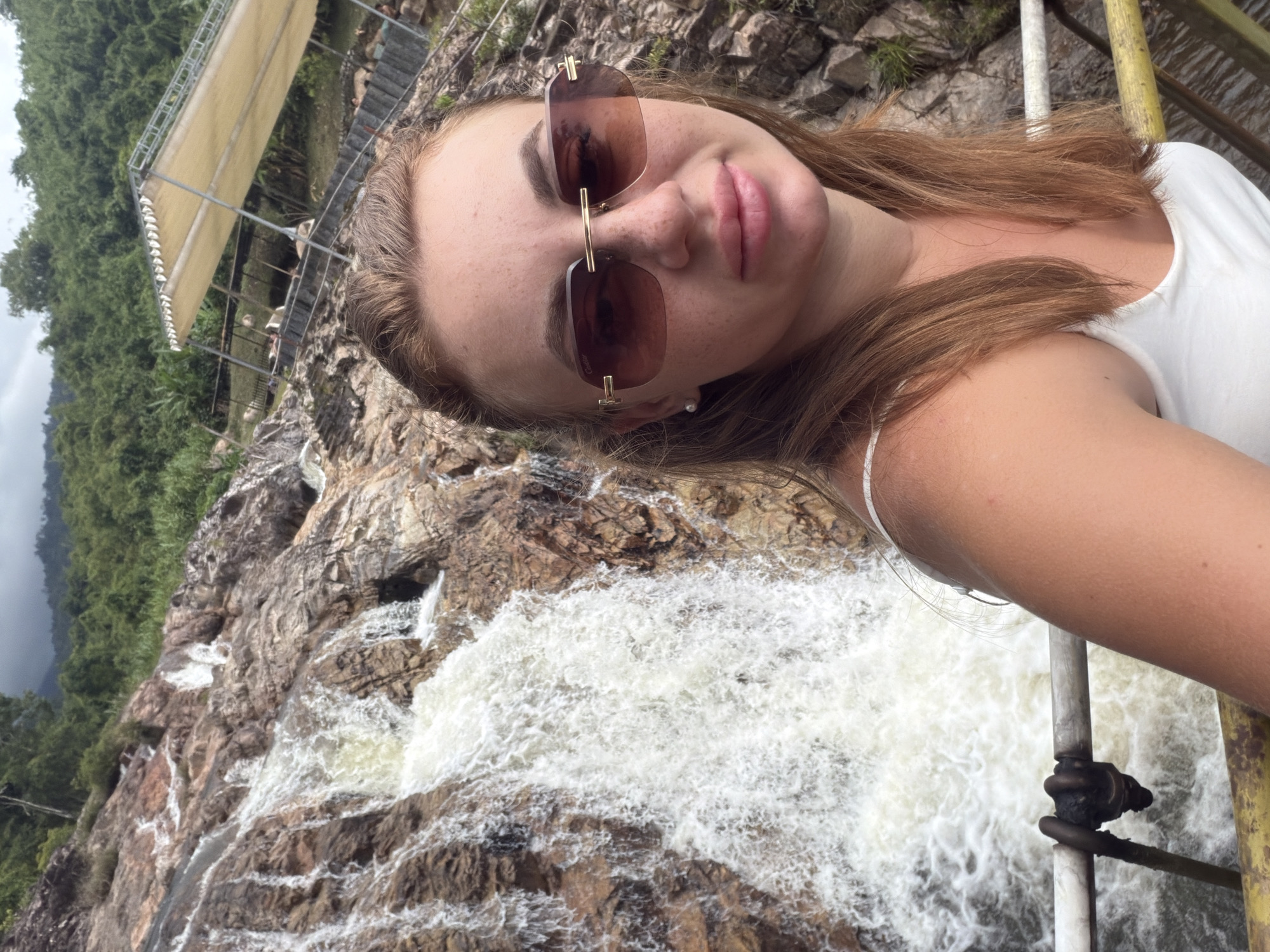 A young woman with light brown hair, wearing a white tank top and oversized sunglasses, is taking a selfie by a waterfall. The waterfall cascades down a rocky slope, with lush green foliage covering the hills in the background. A portion of a metal railing is visible in the foreground, suggesting the presence of a viewing platform or walkway. The woman is smiling slightly at the camera, with a hint of pink lipstick on her lips. The scene appears to be a natural outdoor setting, likely a tourist attraction in a tropical region given the vegetation and weather. The provided context states the location is Khanh Vinh, Vietnam.