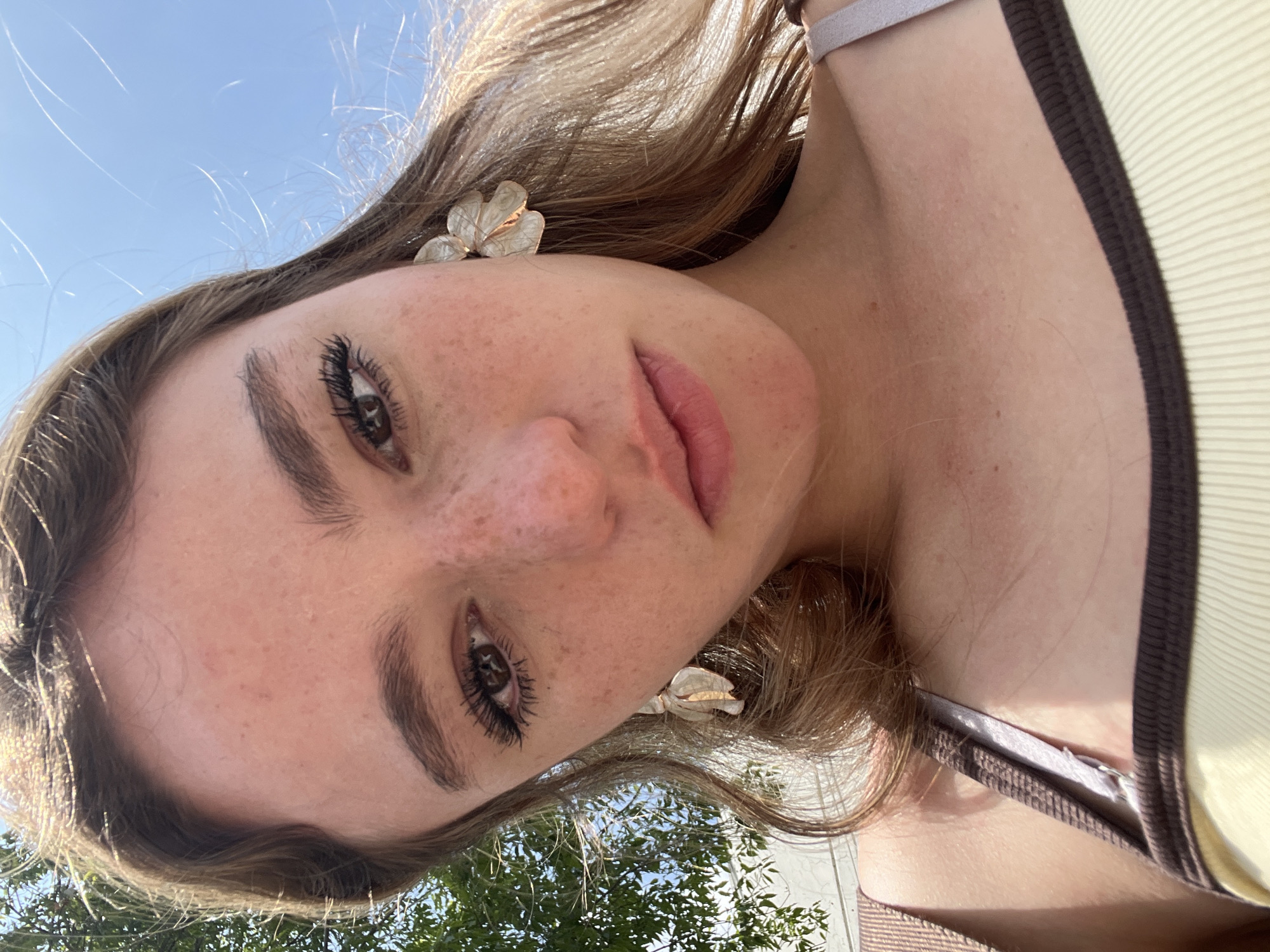 A close-up, slightly angled downward shot depicts a young woman with fair skin and freckles. She has brown hair and is wearing white flower-shaped earrings. Her eyes are brown with long eyelashes, and her lips are full and pink. She is wearing a light-colored top with dark brown trim and a gray strap. The background shows a clear blue sky and the tops of green trees. The location in Puebla, Mexico, is not discernible from the provided image.