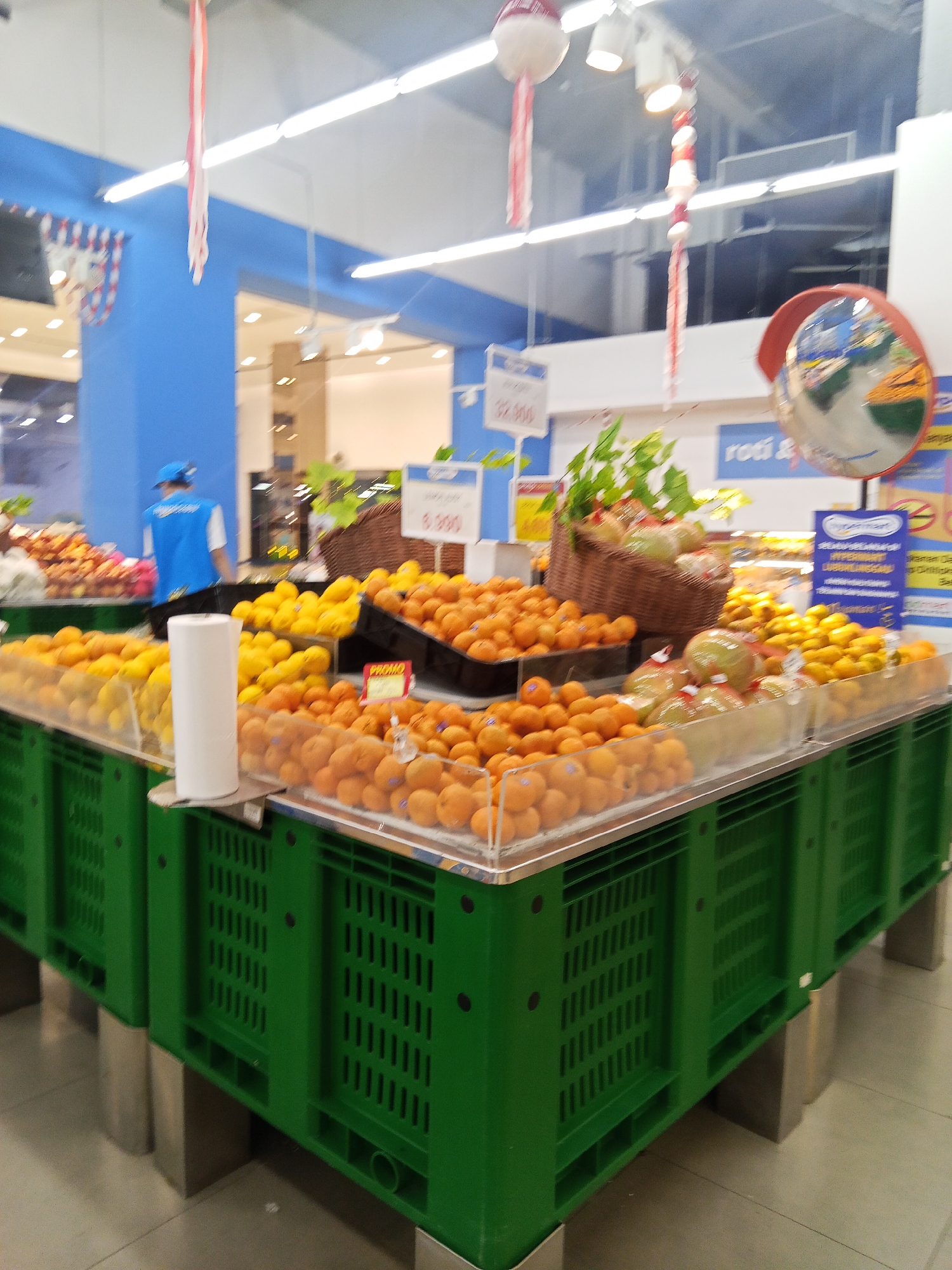 The image shows a supermarket produce section with various fruits displayed in green plastic crates. In the foreground, there are abundant oranges and some lighter-colored fruits, possibly mandarins or tangerines, in clear plastic containers. Behind them, yellow fruits, likely lemons or limes, are visible. A brown basket filled with green produce, possibly melons or gourds, sits to the right. A roll of white paper is placed on a display. In the background, a person wearing a blue uniform and a cap is seen working. Several price tags are visible, one indicating "32.000". Decorative red and white streamers hang from the ceiling. A security mirror is mounted high on the right side of the image. The overall environment is brightly lit with fluorescent lights, characteristic of an indoor retail space. This scene is consistent with the produce section of a supermarket, such as a Hypermart, in Lubuklinggau, Indonesia, given the signage.
