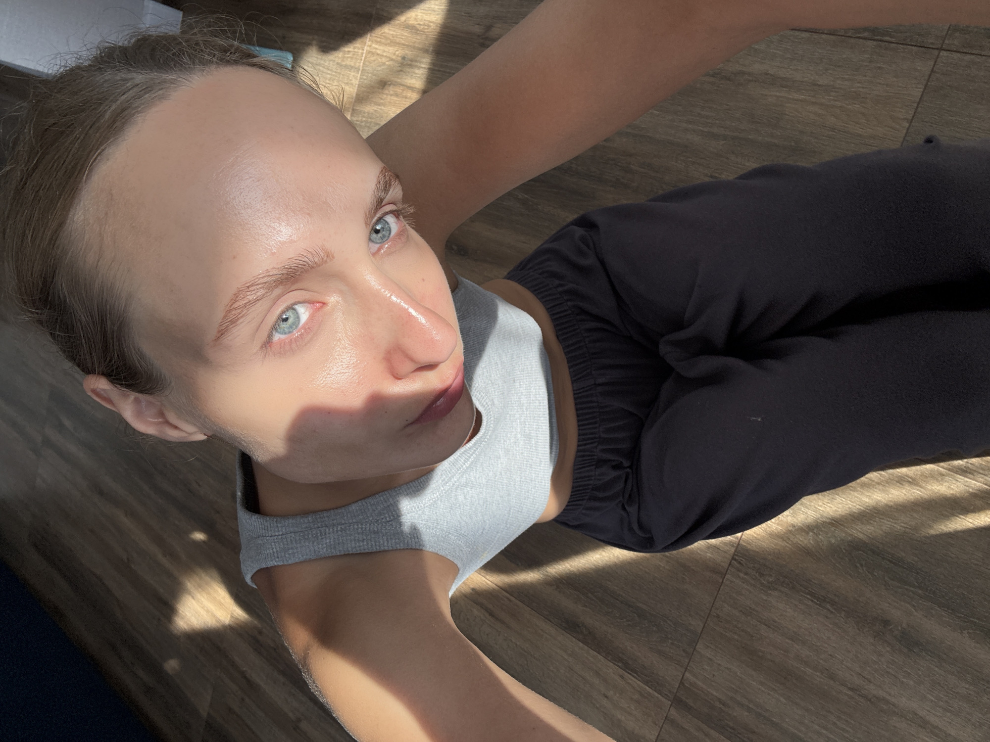 A person is sitting on a wooden floor in what appears to be a home in the city of Podshivalovo, Russia. The person is wearing a gray tank top and dark sweatpants. Sunlight is streaming in through a window, casting shadows across the floor and the person's face. The person has light blue eyes and appears to be looking up at the camera. The background is out of focus, but it seems to be a simple room with wooden flooring.