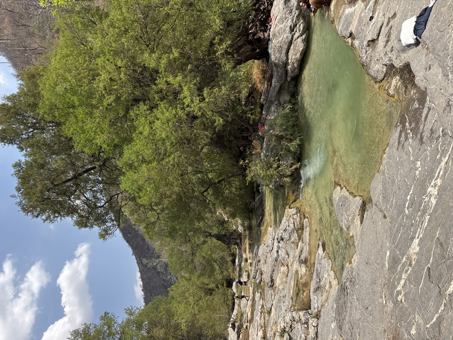 The image depicts a natural riverine environment within San Miguel Atlapulco, Mexico, under a clear blue sky with scattered white cumulus clouds. The foreground consists of large, light-colored stratified rock formations, which form ledges and shallow pools containing clear, greenish-tinted water. In the lower right foreground, a white garment and dark apparel are placed on the rocks.
A stream with light green, flowing water occupies the midground, traversing the rocky terrain and forming small cascades and interconnected pools. On the left bank of the stream, two individuals in dark clothing are walking on the exposed rocks towards the background. On the right bank, a large tree with dense, vibrant green foliage dominates the scene. Beneath this tree, at the water's edge, a person with dark hair, wearing brown attire, is seated on a rock, with their lower legs submerged in the water. Several small, colorful items are visible on the ground near the base of the large tree on the right.
The background features steep, arid-appearing hills or mountains covered with sparse vegetation, including what appear to be cacti and some bare tree trunks. Other green-leaved trees are scattered along the riverbanks extending into the mid-distance. The overall lighting suggests a bright, sunny daytime setting.