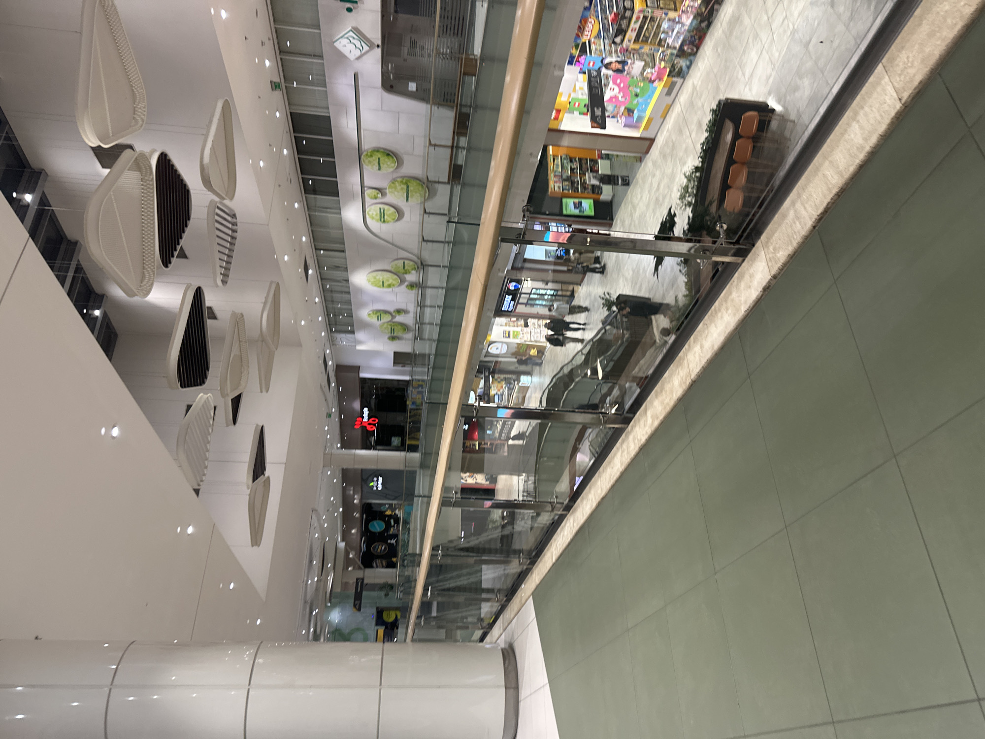 The image shows the interior of a modern shopping mall. From an elevated viewpoint, the scene captures multiple levels with glass railings, revealing shops and walkways below. On the ceiling are decorative hanging fixtures that resemble leaves. There are people walking on different levels of the mall, and various retail outlets are visible, including a toy store. The architecture is characterized by clean lines and a bright, airy atmosphere, which is typical of modern commercial spaces. The location of the mall is in the city of Sofia, Bulgaria.