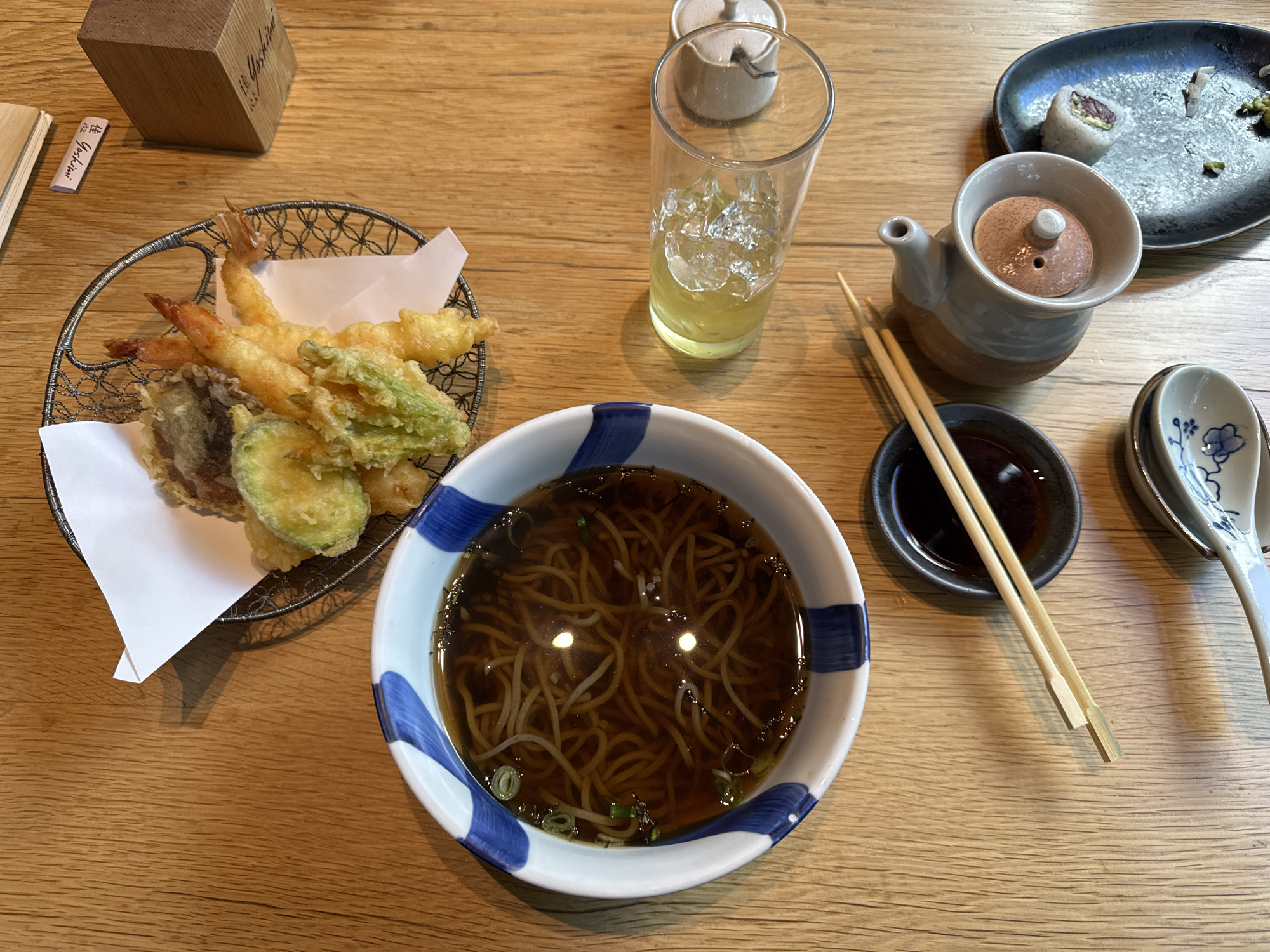 An overhead shot depicts a meal set on a light-colored wooden table at the Japanese restaurant Yoshimi, located within the Hyatt Regency Mexico City hotel in the Polanco neighborhood of Mexico City, Mexico.
In the upper left, a white bowl with a blue striped rim contains soba noodles in a dark broth, garnished with chopped green onions. To its right, a wire mesh basket lined with white paper holds a variety of tempura, including several pieces of shrimp, sliced zucchini, and what appears to be a mushroom and a green leafy vegetable. Above the tempura basket, a small white paper slip and a wooden block, both bearing the name "Yoshimi," are visible.
Centrally, a tall, clear glass filled with ice and a light yellow liquid (likely a beverage) stands to the right of the soba bowl. Below the soba bowl, a small black dish contains a dark liquid, presumably soy sauce, with a pair of light wooden chopsticks resting diagonally across it. Further below, a white ceramic spoon with a blue floral design is positioned. To the right of the chopsticks, a light grey ceramic dispenser with a spouted top and a light brown cap is present. Adjacent to the dispenser, a rectangular dark grey ceramic plate displays remnants of a consumed meal, including one sushi roll with a reddish interior, and scattered small green and light-colored food fragments. Shadows are cast by several objects, indicating an overhead light source.