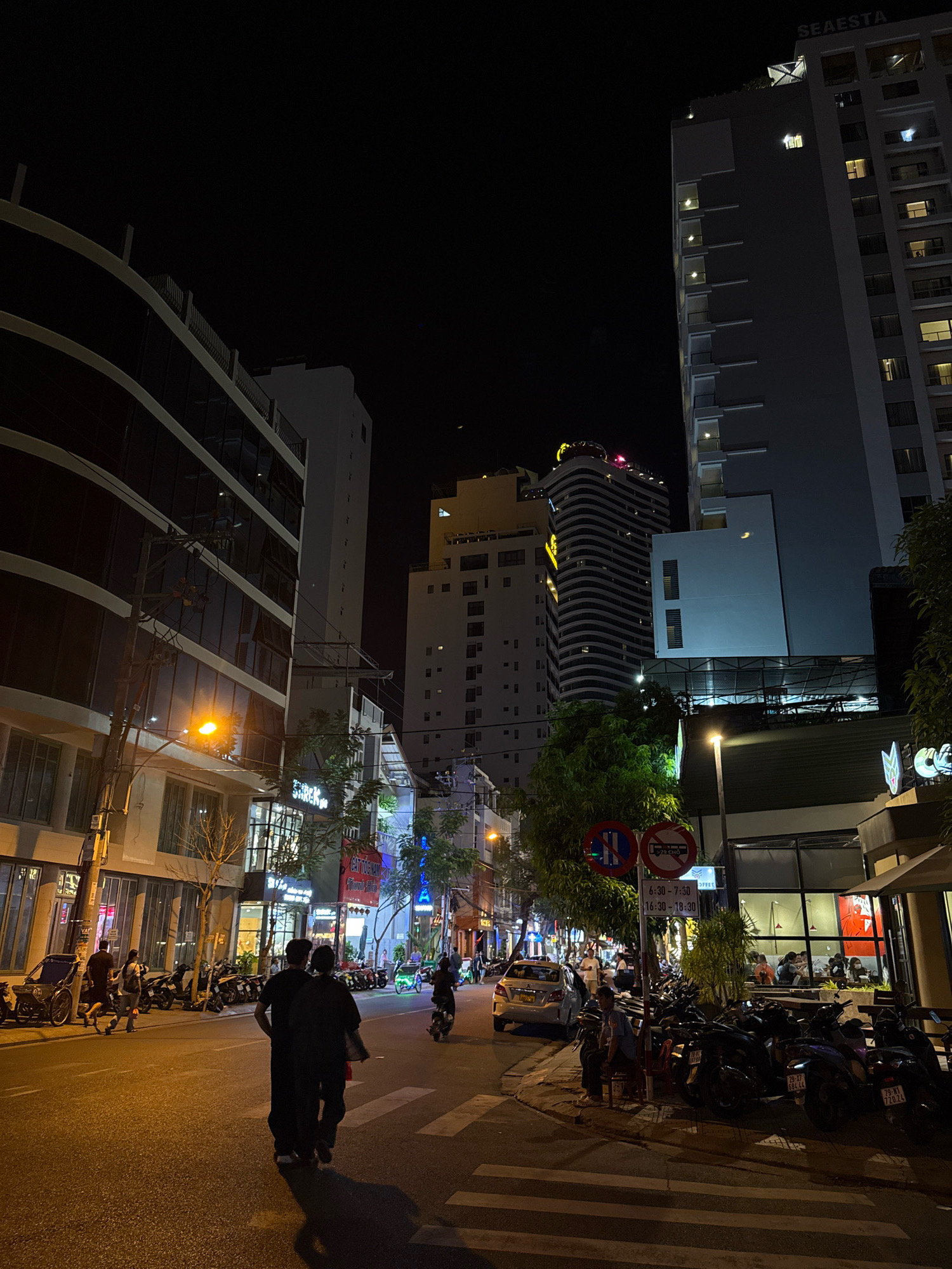 This is a street-level, nighttime photograph taken in Nha Trang, Vietnam, capturing a busy urban scene. The foreground features a paved road with a pedestrian crossing marked by white stripes. Two individuals, seen from behind, are walking away from the camera on the road. To the right, a sidewalk is lined with numerous parked motorcycles. Behind the motorcycles, a building with large glass windows and an awning is visible, with people seated inside. The area is illuminated by streetlights and the lights from the buildings.
In the mid-ground, several buildings of varying heights and architectural styles line the street. One tall building on the right prominently displays the name "SEAEASTA" at its top. Another curved, modern building with illuminated signs stands further down the street. Various shops and businesses with illuminated signs are also present. Trees and utility poles with electrical wires are interspersed throughout the scene.
The background is dominated by the dark night sky, with only the brightly lit buildings providing contrast. The overall atmosphere is that of a lively urban environment at night, with a significant amount of vehicular and pedestrian activity suggested by the parked motorcycles and the people visible in the shops.