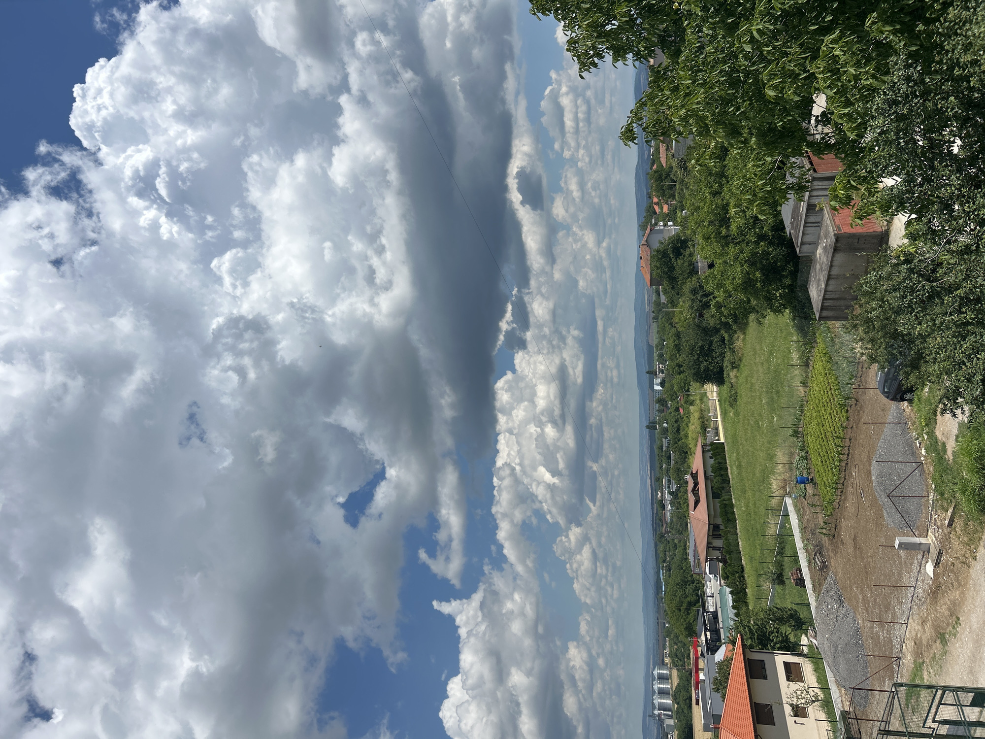 This is a vertical, eye-level photograph taken from an elevated perspective, likely a balcony or rooftop, looking out over a landscape. The dominant feature of the image is the sky, filled with a dramatic array of white and grey clouds against a bright blue background. Some clouds are fluffy and white, catching the sunlight, while others are darker and more ominous, suggesting the possibility of changing weather.
Below the sky, the scene transitions to a residential area with houses featuring red-tiled roofs interspersed with lush green trees. A neatly manicured grassy field with rows of plants, possibly a vineyard or garden, is visible. A gravel path winds through the area, bordered by a fence. In the distance, a town or city can be seen nestled among rolling hills under a hazy sky.
There are no people or specific activities explicitly depicted. The overall impression is one of a serene, rural or semi-rural setting under a dynamic sky. The time of day appears to be daytime, given the bright illumination and the blue sky peeking through the clouds. There is no visible text in the image. The elevated viewpoint and the expanse of sky and landscape create a sense of openness and breadth.