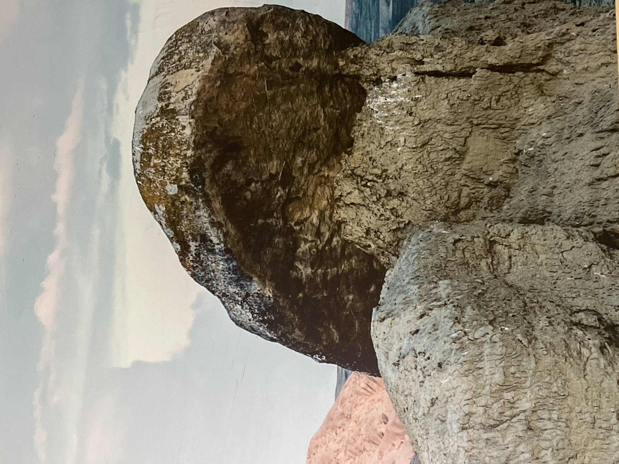 The image displays a close-up view of large, weathered rock formations against a hazy sky with clouds. In the foreground, a lighter-colored, rough-textured rock is visible. Behind it, a larger, darker rock formation with patches of moss and lichen dominates the center of the frame. To the left, a pinkish, textured geological formation is partially visible. The background consists of a blurred expanse of water and a sky with soft, diffused lighting. The overall scene suggests a natural, outdoor environment, likely a coastal or rocky area. The specific location of Sabail Raion, Azerbaijan is not discernible from the visual elements within the image. There are no people, animals, or man-made objects depicted.