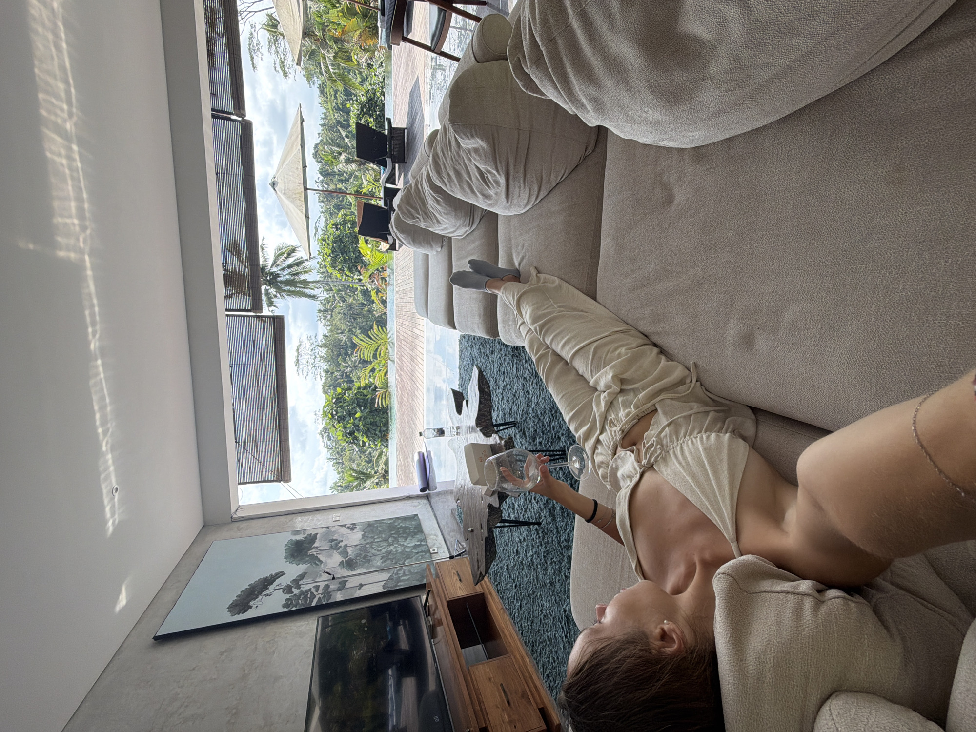 The image shows a woman lying on a couch, holding an empty wine glass. She is wearing a light-colored top and pants. A television and artwork are visible in the background, along with a large window that looks out onto a tropical landscape. The location is likely in Petulu, Indonesia, based on the style of the architecture and the visible landscape.