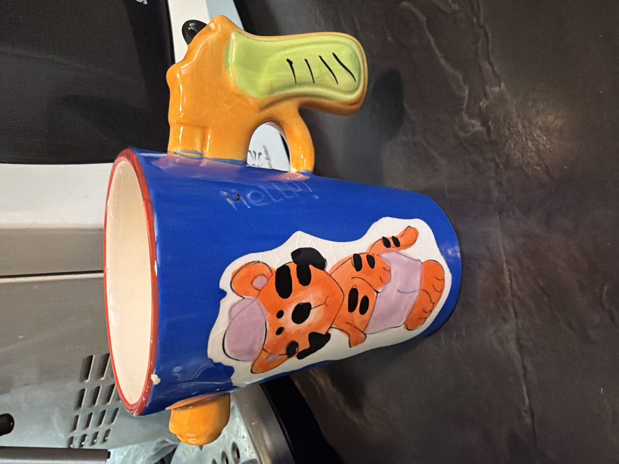 The image shows a blue mug with a cartoon depiction of Tigger from Winnie the Pooh wearing a diaper, lying on its side. The mug handle is orange and green. The mug appears to be resting on a dark gray surface, possibly a leather or synthetic material. A white object with ventilation slots is partially visible in the background. There is no information to suggest the image was taken in Pleven, Bulgaria.